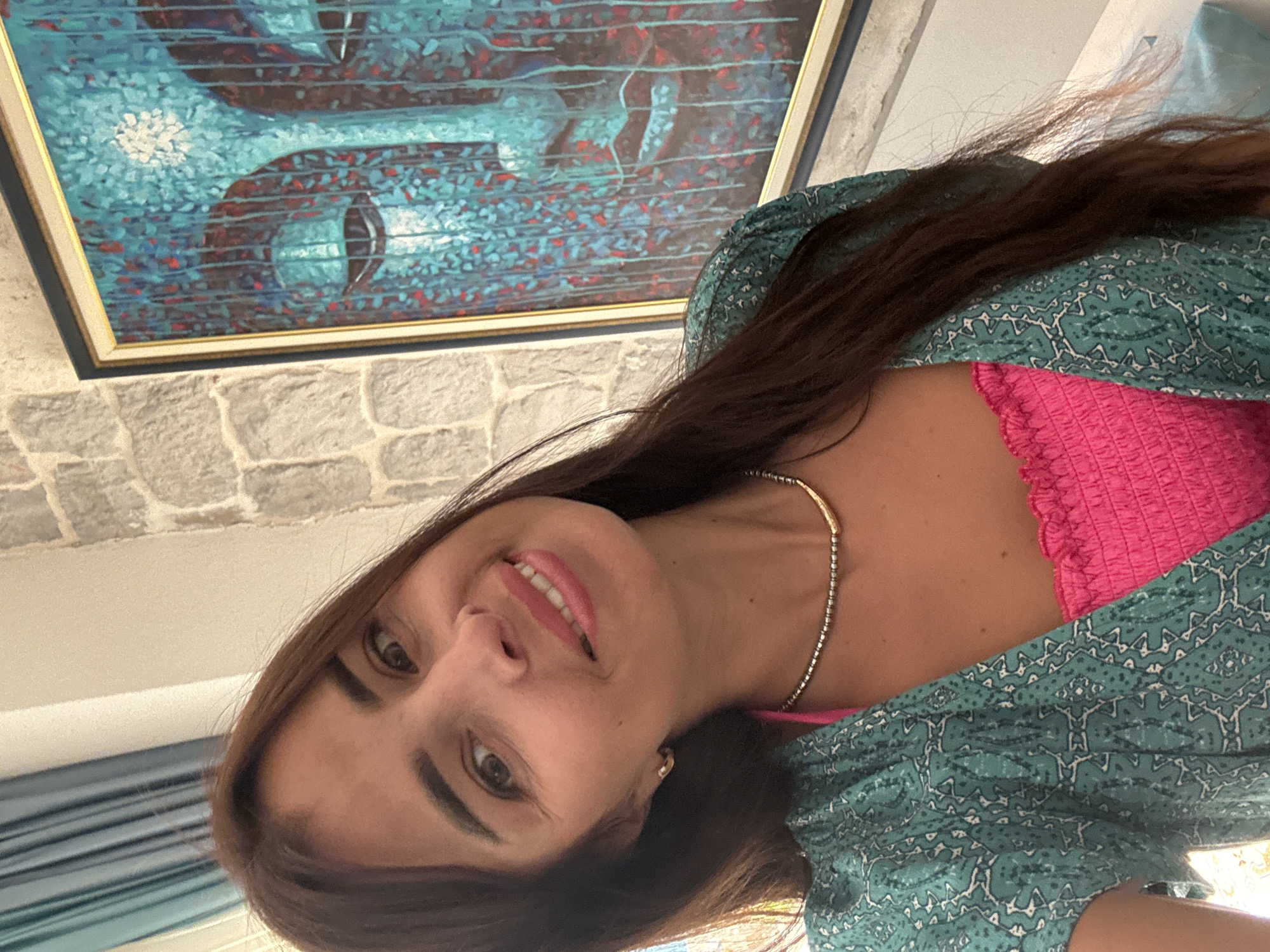 An upper-body, medium shot shows a woman with long, dark brown hair smiling at the camera. She is wearing a bright pink ruffled top and a teal patterned open-front blouse. A silver beaded necklace with a gold bar is around her neck. In the background, a framed painting of a blue and red abstract Buddha face hangs on a stone wall. To the left of the painting and woman are turquoise curtains.