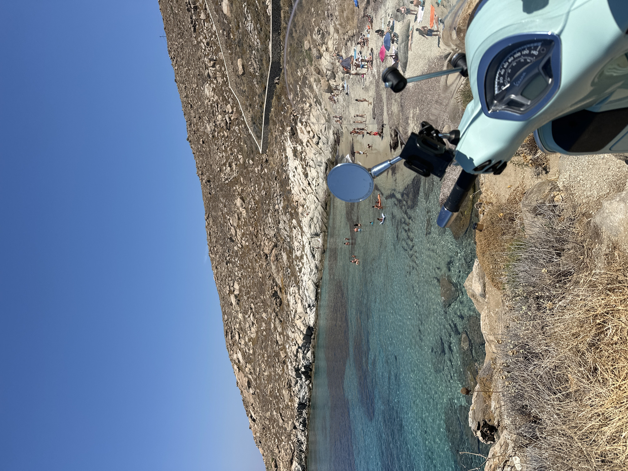 The image depicts a scenic view of a beach at Agios Ioannis Diakoftis, Greece, framed by a light blue scooter in the foreground. The scooter's handlebars and dashboard are visible, with a mirror reflecting the sky and a mount for a device. Beyond the scooter, a crescent-shaped beach with light-colored sand is populated by numerous beachgoers. Some individuals are swimming or wading in the clear, turquoise water, while others are sunbathing on the sand, with several beach umbrellas providing shade. The coastline is characterized by rocky hills dotted with sparse vegetation. The water is calm and shallow near the shore, gradually deepening to a darker blue further out. The sky above is a clear, cloudless blue. A stone wall with a winding path is visible on the hillside. The overall impression is one of a sunny, relaxed day at a popular beach destination.