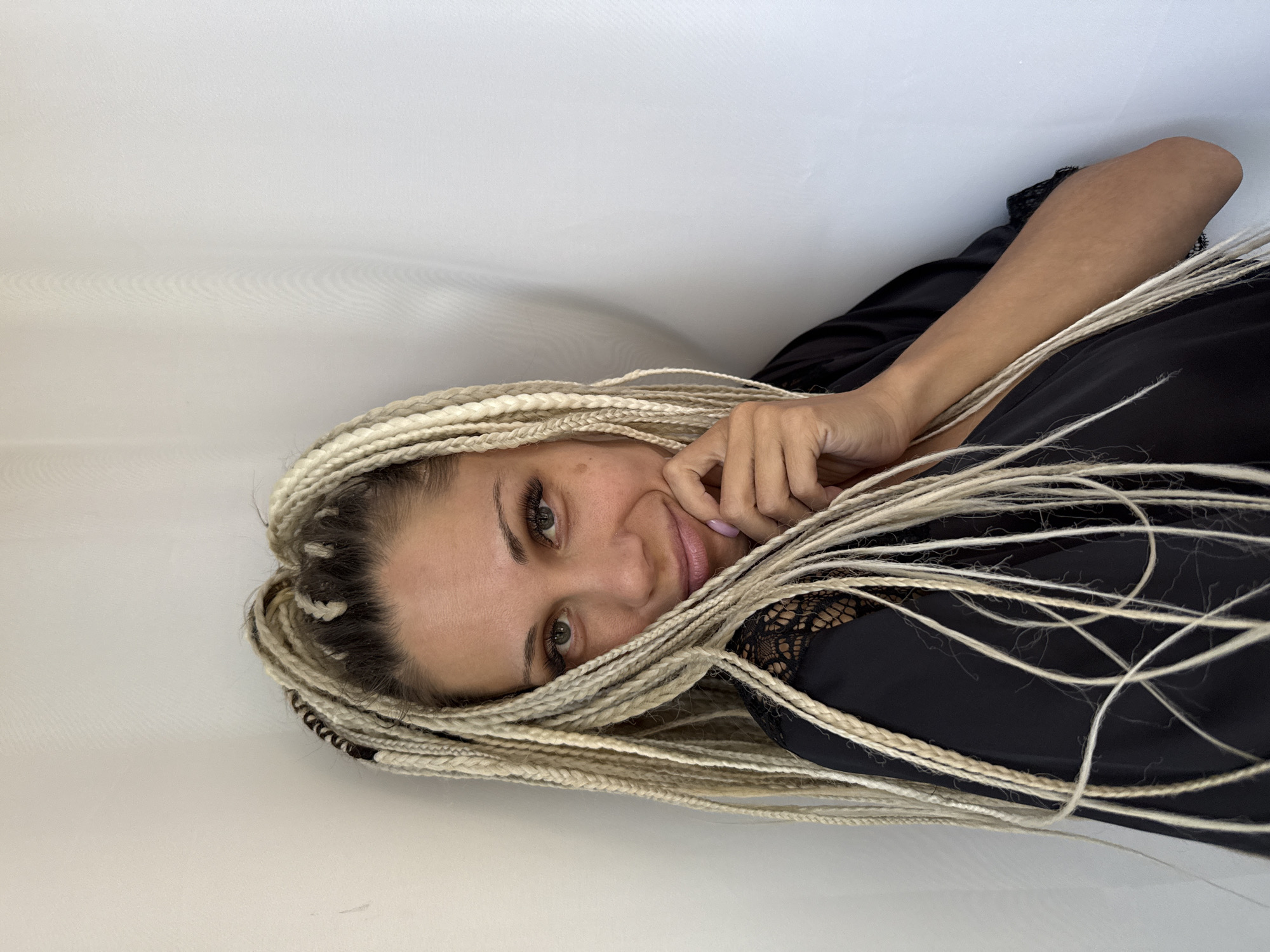 The image shows a portrait of a woman with blonde braided hair against a plain white background. She is wearing a black garment with lace detailing. The woman is looking towards the camera and has her hand near her face. The lighting is bright, suggesting an outdoor or well-lit indoor setting. The location is specified as Rusanovo, Russia.