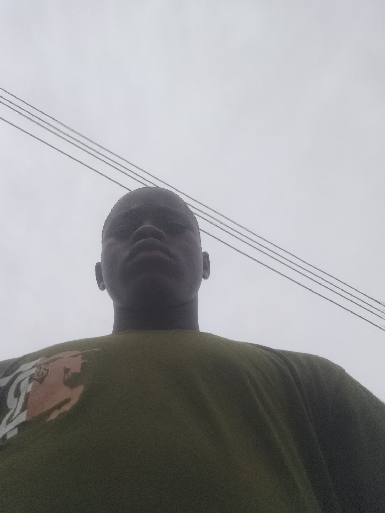 A low-angle shot shows a dark-skinned person wearing a green shirt with a design, possibly standing outdoors. Above them, against an overcast sky, are several parallel power lines. The location is assumed to be in Epe, Nigeria, based on the prompt.