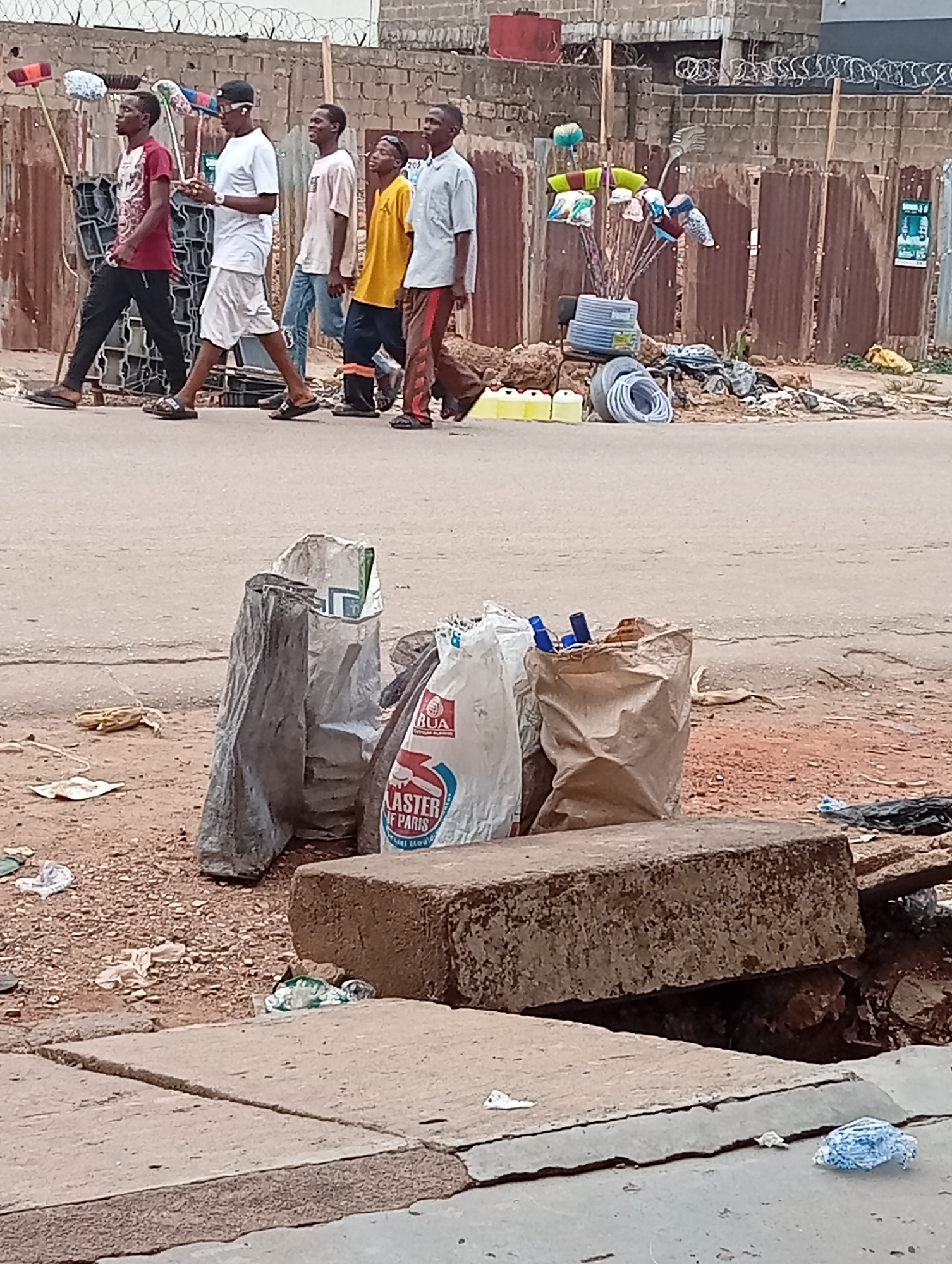 The image depicts a street scene in Maiburiji, Nigeria. In the foreground, a partially collapsed sidewalk is visible, exposing the ground beneath. Three large sacks, one bearing the inscription "BUA, MASTER OF PARIS, rual Medic," sit on the uneven ground near the sidewalk. Litter is scattered nearby.
In the background, five men are walking along the road. The first man is wearing a red shirt and black pants, carrying cleaning supplies. The second man is wearing a white shirt and shorts. The third is wearing a pink shirt and jeans. The fourth is wearing a yellow shirt. The fifth is wearing a light blue shirt and patterned pants.
Behind the men, a corrugated metal fence stands, topped with barbed wire. A display of cleaning supplies, likely for sale, is set up on the roadside. Other objects such as yellow jerrycans and white PVC hoses are also visible. The street itself is paved but shows signs of wear and tear.