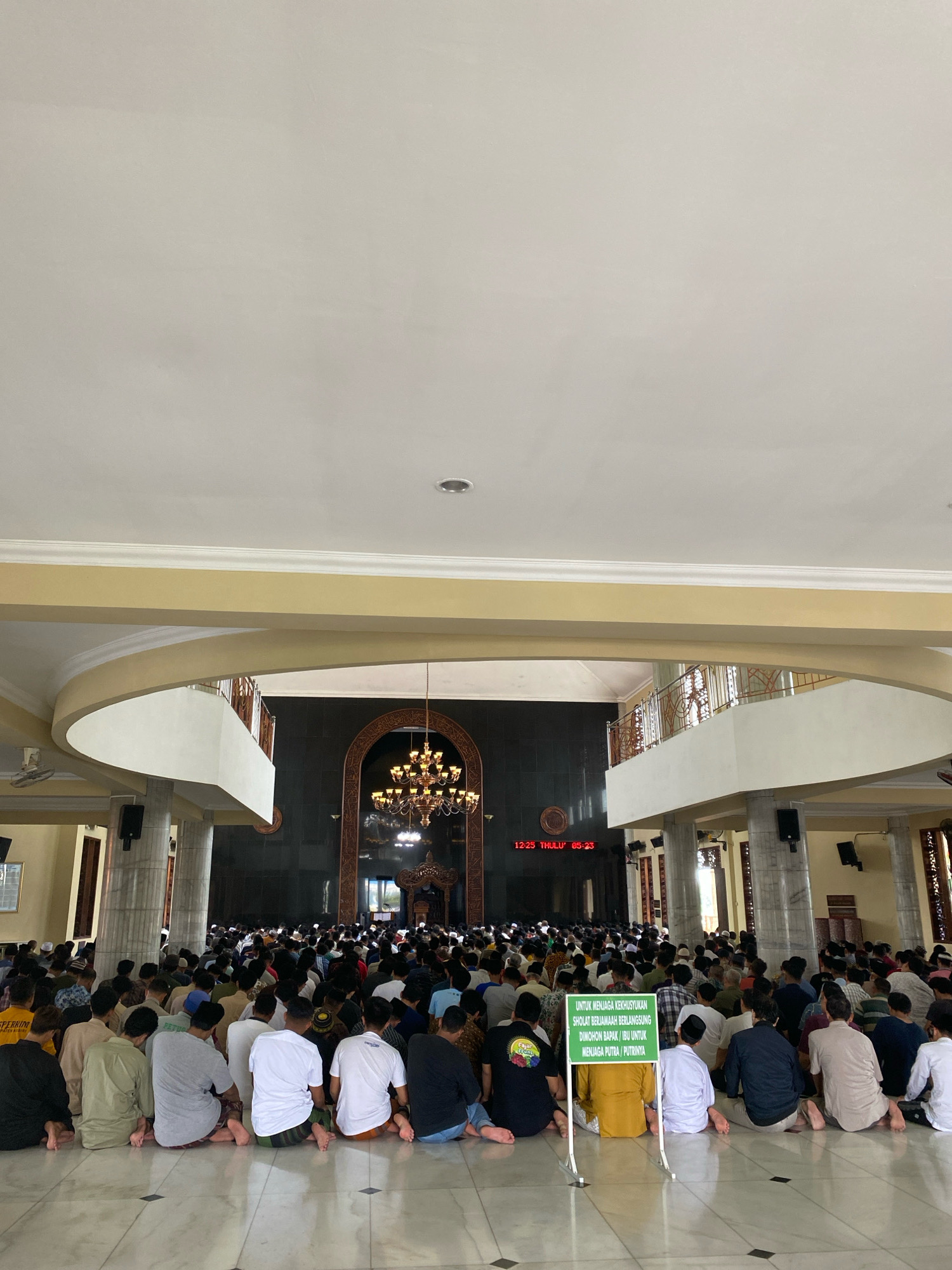 The image depicts a large group of men seated on the floor of a mosque, facing away from the camera, during prayer. The mosque features high ceilings with ornate chandeliers and decorative arches. A digital clock displays "12:25 THULUTHI 05:23" on a dark wall. A green sign with white text is prominently displayed in the foreground, reading: "UNTUK MENJAGA KEKHUSYUKAN SHOLAT BERJAMAAH BERLANGSUNG DIMOHON BAPAK/IBU UNTUK MENJAGA PUTRA/PUTRINYA" which translates to "TO MAINTAIN THE SOLEMNITY OF THE CONGREGATIONAL PRAYER, WE REQUEST FATHERS/MOTHERS TO SUPERVISE THEIR SONS/DAUGHTERS." The floor is tiled in a marble pattern, and the environment appears to be indoors, with pillars and arched entrances visible in the background. The men are dressed in various casual attire, with some wearing traditional Islamic clothing. The overall scene conveys a sense of communal worship and order.