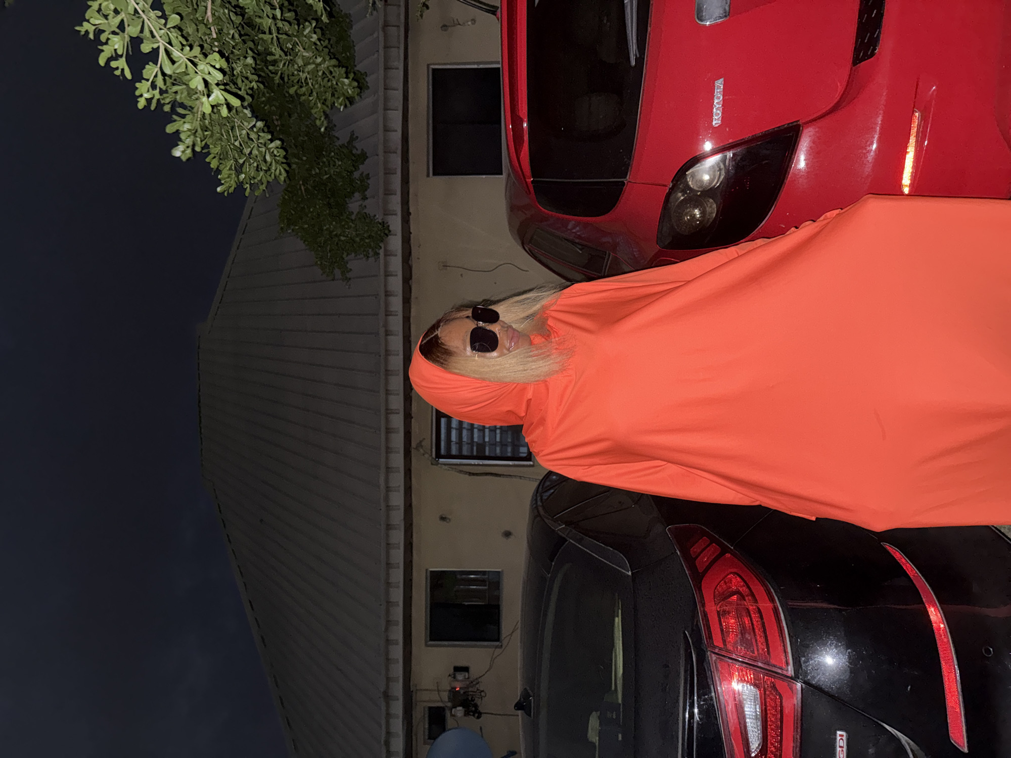 A woman stands in front of a residential building in Ado, Nigeria. She is wearing an orange hooded garment and sunglasses, and her blonde hair is visible. Two cars, one black and one red, are parked behind her. The black car is to her left, and the red car is to her right. The building behind her has a corrugated metal roof and several windows. A tree with green leaves is visible to the right of the building. The sky is dark, suggesting it is nighttime. The scene appears to be a residential area.