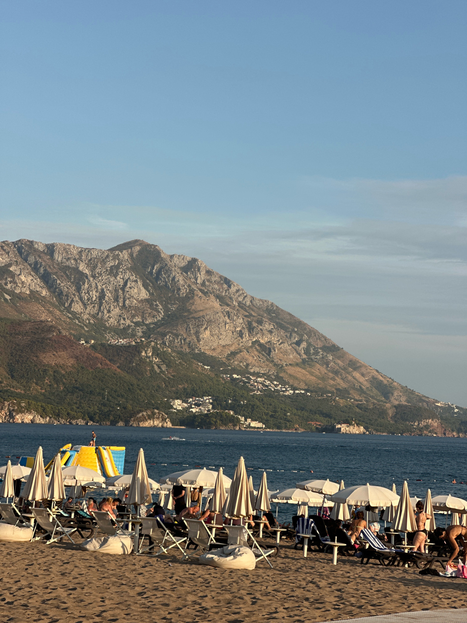 The image depicts a scenic view of a beach in Boreti, Montenegro. In the foreground, a sandy beach is lined with numerous white umbrellas and lounge chairs, some occupied by sunbathers. Beyond the beach, the calm, blue waters of the Adriatic Sea stretch towards a majestic mountain range. The mountains are rugged and covered with sparse vegetation and scattered buildings, indicating a coastal town nestled on the slopes. In the middle ground, a large, inflatable water slide, predominantly yellow and blue, stands prominently in the sea, with a person visible at the top, suggesting recreational activity. The sky is a clear, soft blue with a few wispy clouds, and the overall lighting suggests it is late afternoon or early evening. The scene is tranquil, with a mix of natural beauty and leisure activities.