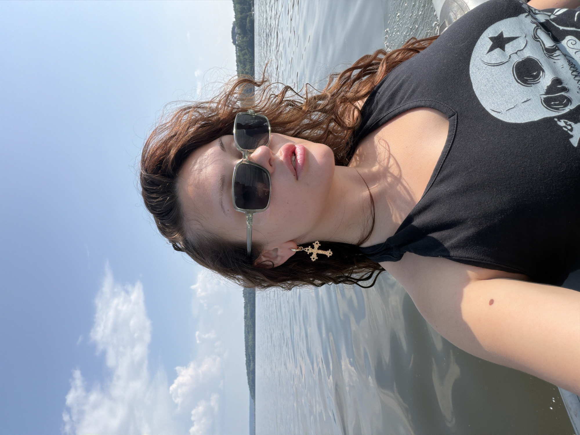A woman wearing sunglasses and a black tank top with a skull graphic is on a boat. She has curly brown hair and is wearing a gold cross earring. The boat is on a body of water, with a tree-lined shore in the background. The sky is blue with white clouds. The location is the city of Sylva, Russia.