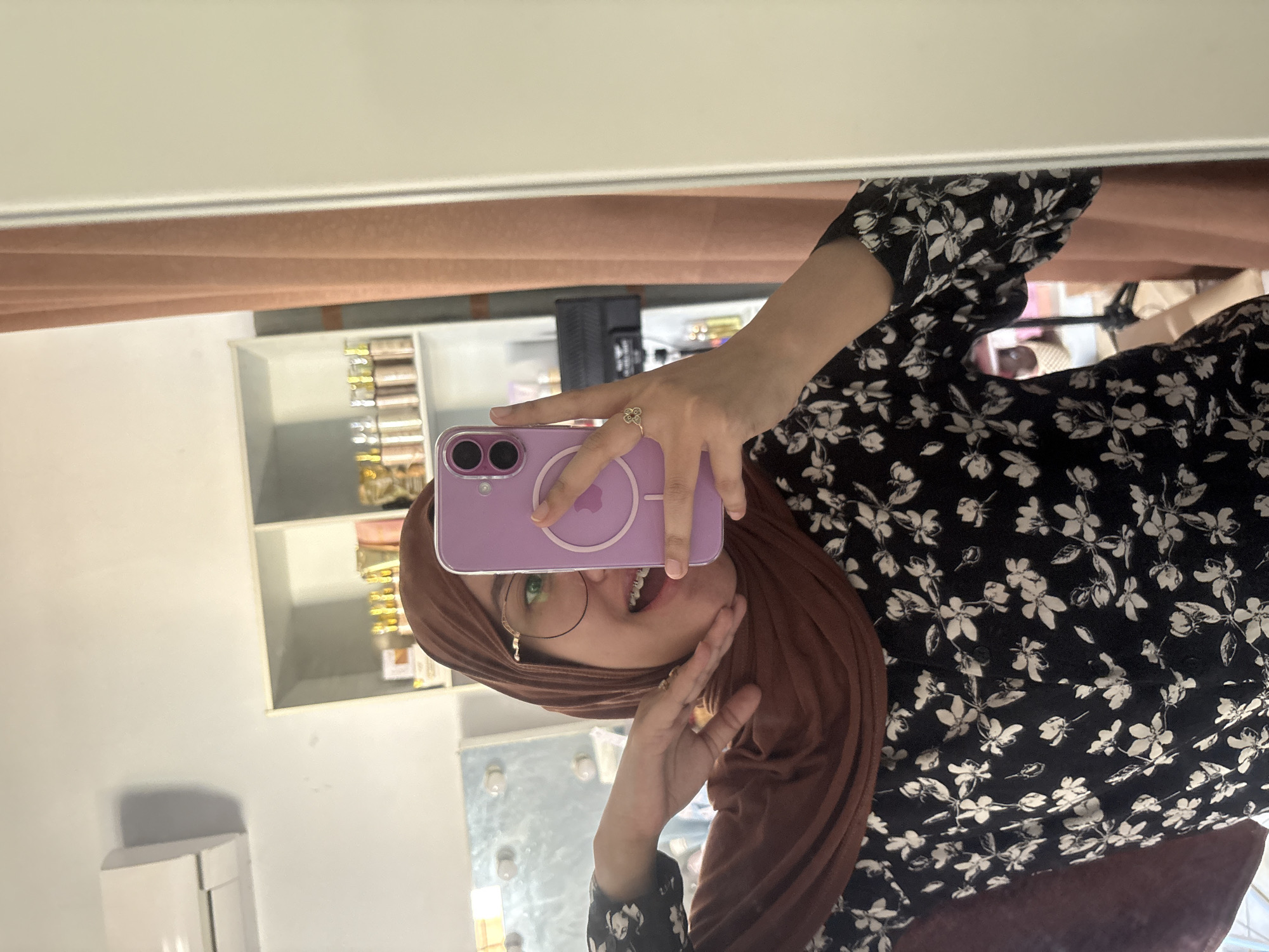 A young woman in a brown hijab and floral-patterned dress takes a selfie in a mirror. She holds a purple iPhone in front of her face. Her left hand is near her cheek, and a ring is visible on one finger. A shelf is seen in the background, possibly in the city of Gadingrejo, Indonesia.