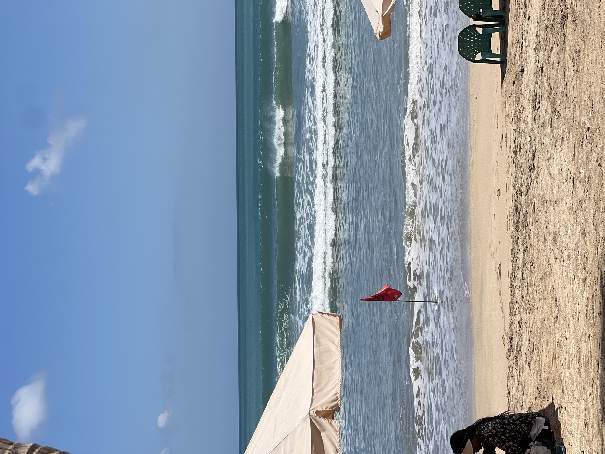The image depicts a beach scene in Kuta, Indonesia, on a sunny day. The foreground is dominated by sand, with a person sitting on the left side, facing away from the camera. They are wearing a dark floral patterned top and a visor. Beside them are some bags. In the middle ground, a red flag is planted in the sand, with waves crashing on the shore. Two green plastic chairs are visible on the right side of the sand. In the background, the ocean stretches out to the horizon, with clear blue skies above, featuring a few scattered clouds. Two beach umbrellas are partially visible, one on the left and one on the right, providing shade over the sand. The waves are moderate, suggesting conditions suitable for recreational activities.