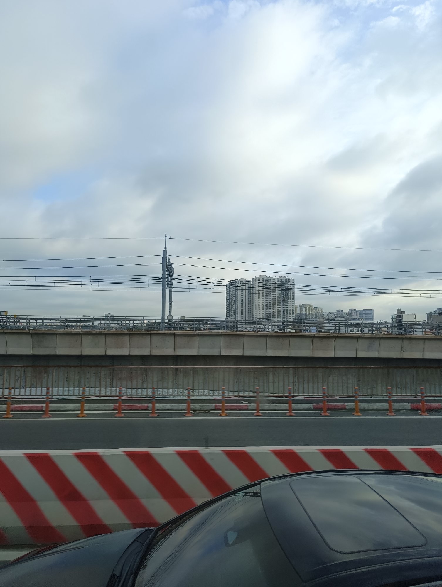 A wide shot captures a view from a moving vehicle, focusing on an elevated highway or railway overpass against a cloudy sky. In the foreground, the hood and sunroof of a dark car are visible. Beyond the car, a concrete barrier with red and white diagonal stripes marks the edge of the roadway. The overpass itself is a substantial concrete structure. Above the overpass, a series of electrical wires are strung between poles, with one tall pole featuring communication equipment. In the background, several multi-story buildings, indicative of an urban or suburban environment, are visible. The overall scene suggests travel through a developed area with significant infrastructure. There are no people or animals depicted in the image. The location is identified as Nakhabino, Russia.