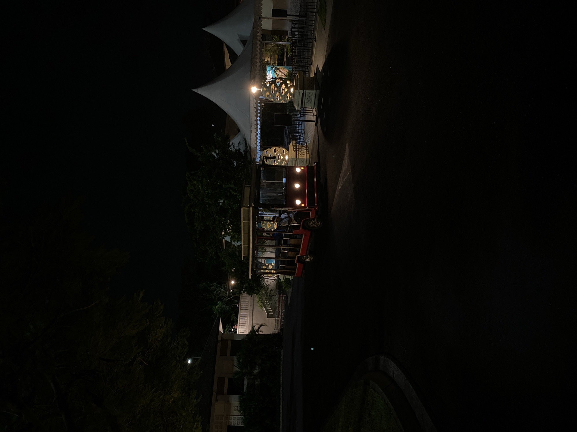 In the city of Kuta Selatan, Indonesia, at night, a red and silver tram navigates a paved road in front of a resort. The tram is open-air, with a driver visible at the front. The resort features white, conical tents and two decorative stone pillars with intricate cutouts, illuminated by internal lighting. To the left, a building with white walls and a balcony can be seen, surrounded by trees and foliage. The scene is dimly lit, with artificial lights from the resort and the tram providing the primary illumination.