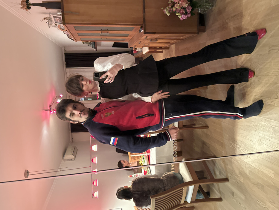 A mirrored reflection showcases two individuals. One, in a red and blue track jacket with the Russian flag, stands behind a person in a white blouse and black corset-like top with matching black pants. The person in the foreground holds a smartphone, presumably taking the picture. The reflection also shows a dining table with people seated, and a furry object on a chair. Red pendant lights hang from the ceiling, and a cabinet with flowers on top stands in the background.
