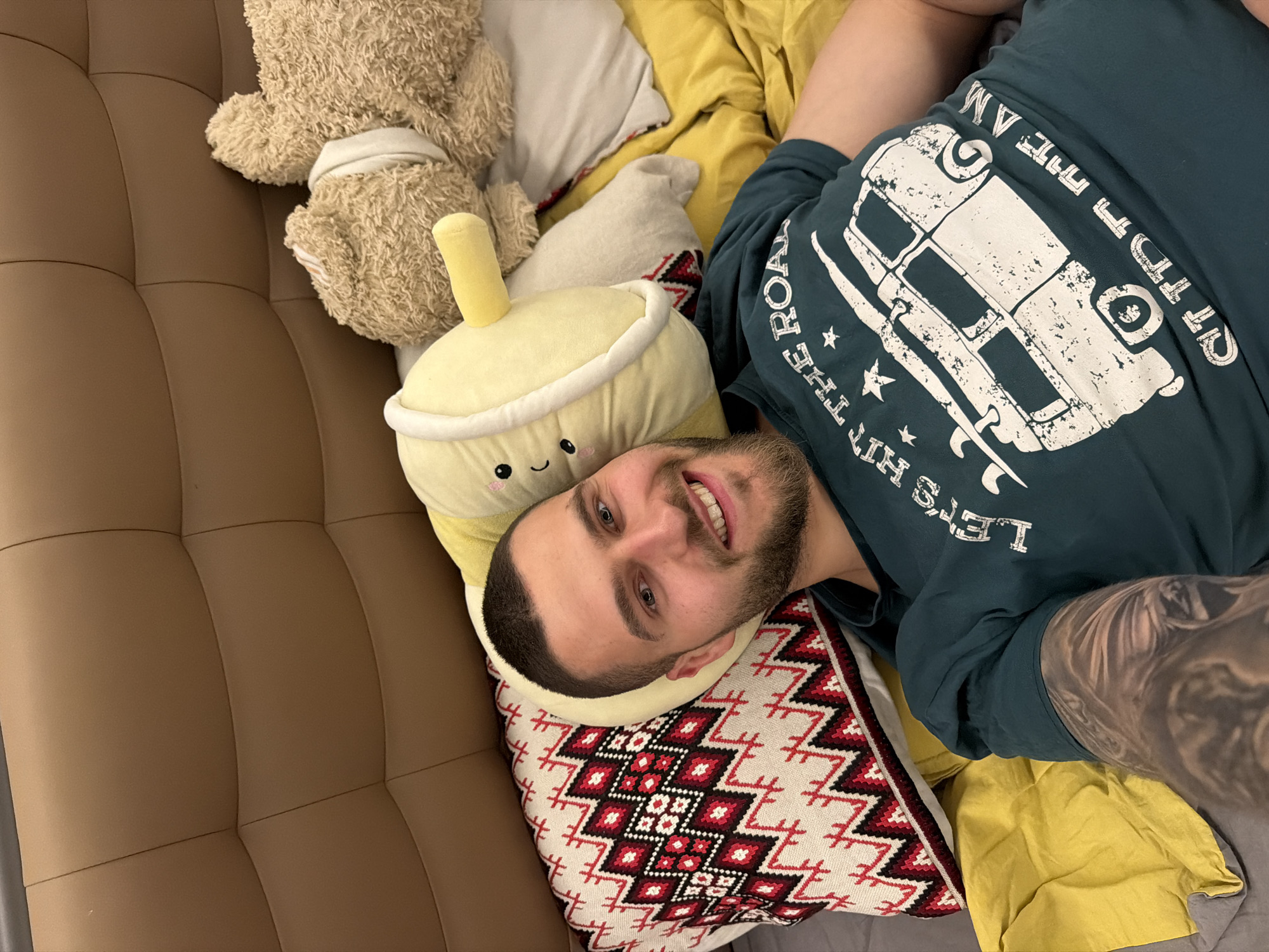 A man is depicted lying down indoors, smiling directly at the camera. He has short dark hair, a beard, and light-colored eyes. He is wearing a dark teal t-shirt featuring a large white graphic print of a vintage camper van or microbus. Visible text on the shirt reads "LET'S HIT THE ROAD" along the bottom and partially "OFF THE AM" along the top, with the remainder obscured. A portion of his right arm shows a large, intricate tattoo.
He rests his head on a white pillow adorned with a red and black geometric, possibly ethnic, pattern. This pillow is placed on what appears to be a yellow blanket or duvet. Beside him, on his left, is a soft yellow boba tea-shaped plush toy with an embroidered smiling face. Further in the background, a light brown teddy bear plush is visible. The man and the plush toys are set against a brown upholstered surface, likely a sofa or headboard, characterized by a quilted pattern. The scene conveys a relaxed and comfortable atmosphere, and the man's expression suggests happiness or contentment. The photograph is situated in Sofia, Bulgaria.