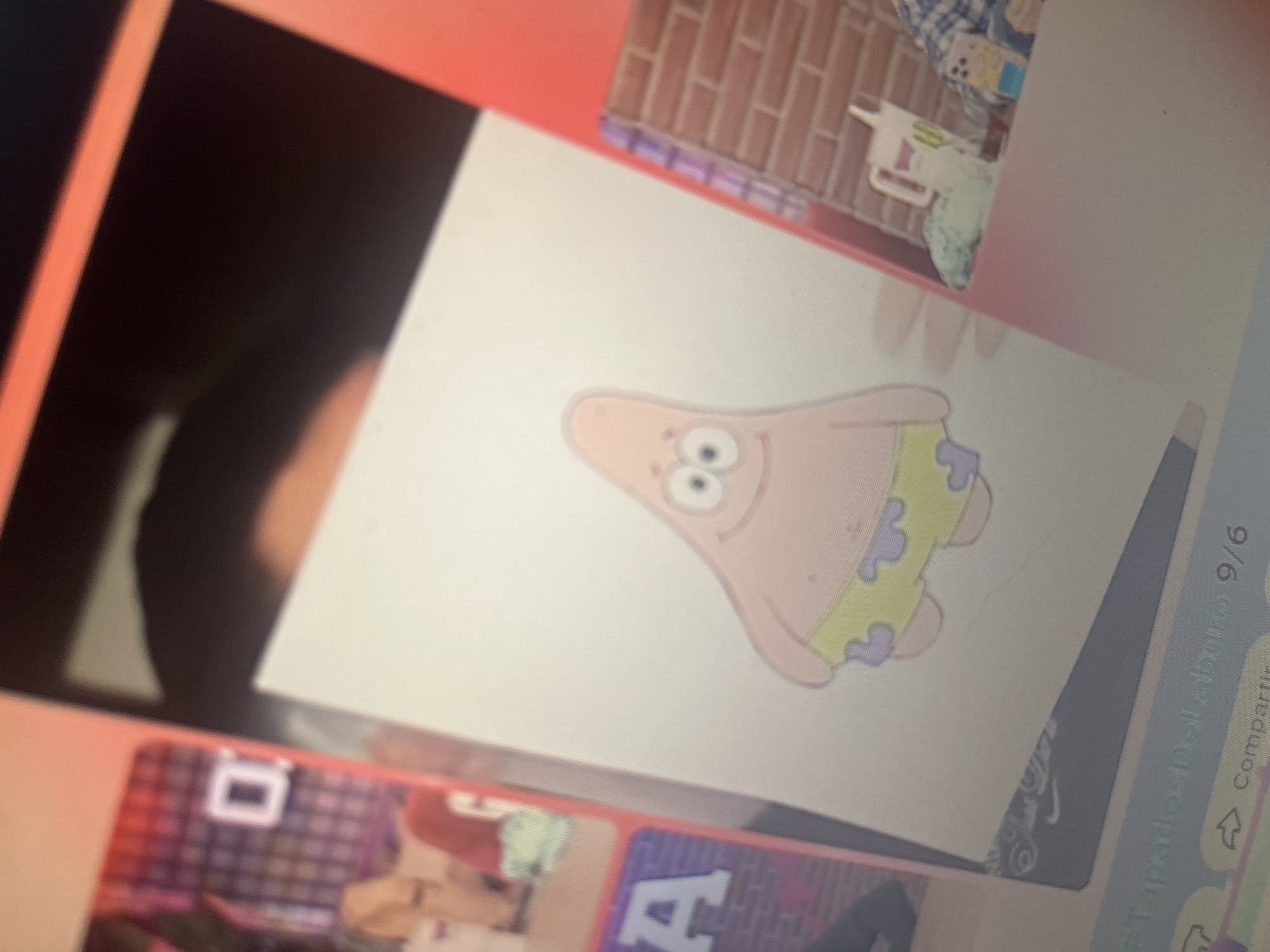 The image depicts a drawing of Patrick Star from SpongeBob SquarePants on a piece of white paper. Patrick is depicted with his characteristic pink body, large eyes, a smiling mouth, and wearing green shorts with purple polka dots. The drawing is placed on a surface, likely a table, with other elements visible in the background. To the left of the drawing, a portion of a screen or display is visible, showing what appears to be a background with a grid pattern and a white "0" or "D". Further to the left, a purple sign with a white letter "A" is visible. To the right of the drawing, a brick wall texture is evident, with a white pitcher-like object and a colorful bottle on a surface in front of it. The bottom of the image shows a partial view of a device screen with text that includes "#TapadosDelaburo 9/6" and "Compartir". The scene is illuminated by an overhead light source, casting subtle shadows. There is no clear indication or context within the image that places this scene in the city of Santa Ana, Venezuela.