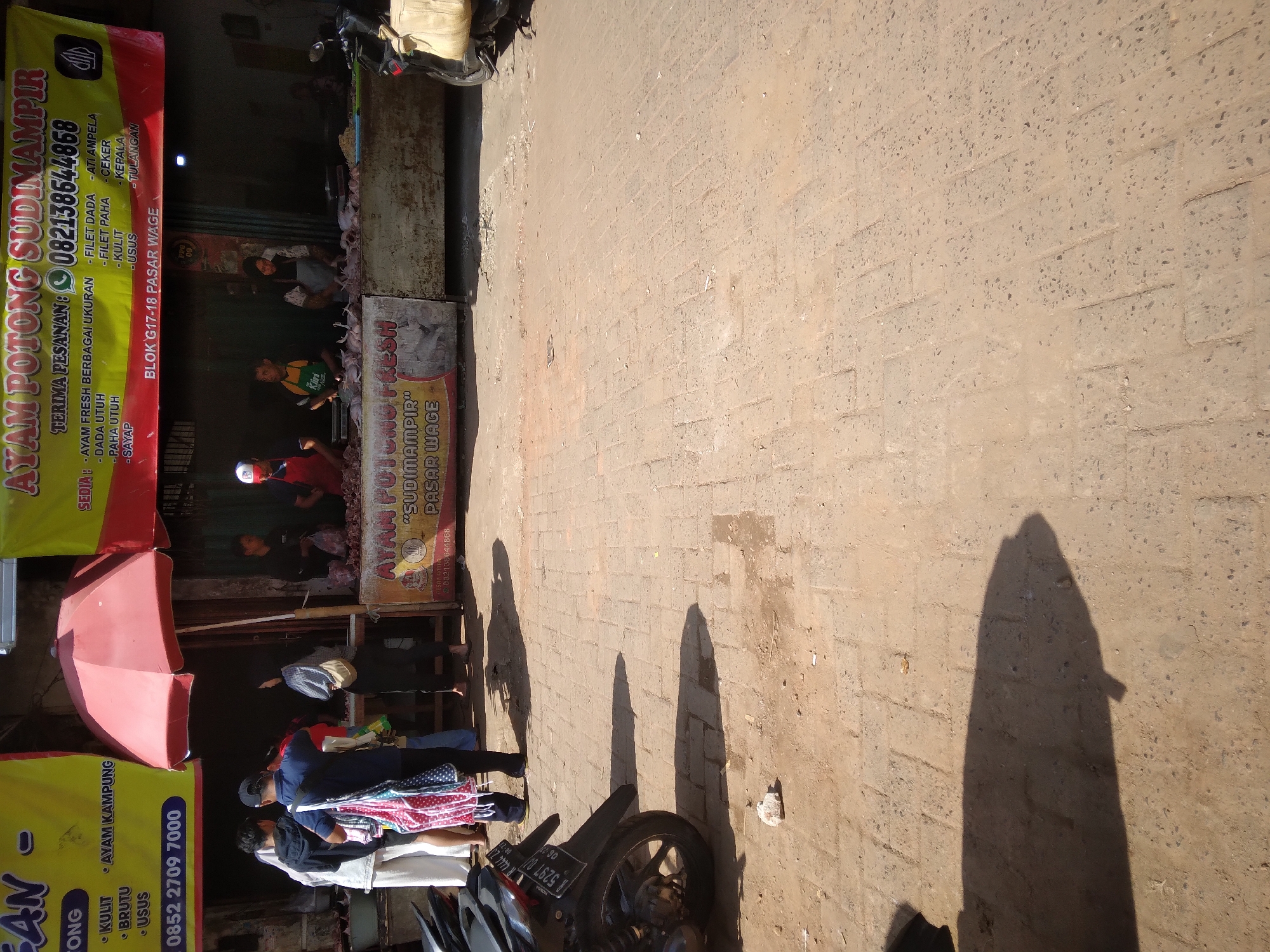 A street market scene in Purwokerto, Indonesia is depicted, likely on a sunny day given the strong shadows. The foreground shows a brick-paved ground with a motorcycle on the left, its license plate reading "R 5297 DJ". In the background, a food stall with a red umbrella is set up, selling chicken. Several individuals are visible, some appearing to be customers and others working at the stall. Banners with text in Indonesian are displayed above the stall, advertising "AYAM POTONG SUDIMAMPIR" and providing a phone number. The overall environment suggests a bustling local marketplace.