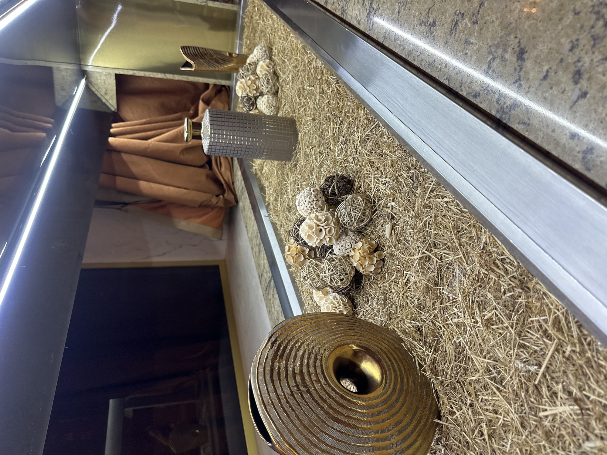The image shows a close-up, eye-level view of a decorative display on a counter in Estosadok, Russia. The display is arranged in a long, rectangular metallic trough filled with dried straw. On the left side of the trough, a large, golden, circular vase with concentric ridges is prominently featured. To the right of the vase, scattered throughout the straw, are several clusters of small, decorative balls made from natural materials like rattan and dried flowers in beige and brown tones. Further to the right, a tall, cylindrical, grey patterned vase stands upright. Behind the display, draped brown fabric hangs against a wall. The counter is a speckled granite material. Overhead, bright white LED lights illuminate the scene, reflecting on the metallic surfaces and the granite. The overall impression is one of interior decoration, possibly in a hotel lobby or a retail space.