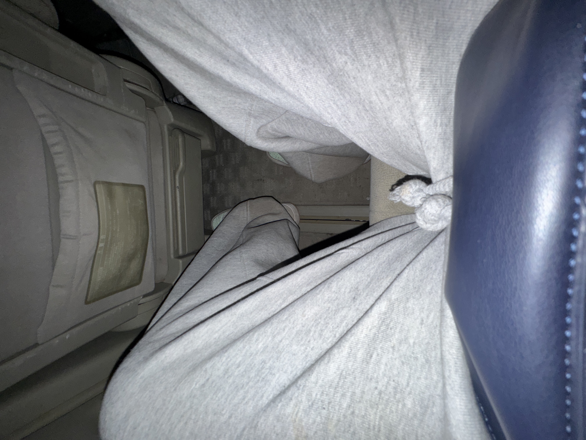 A close-up shot, indoors, shows a person seated on what appears to be an airplane. The person is wearing grey sweatpants with a drawstring. A blue leather seat is visible to the right, and the plane's wall panel is to the left. The location is unknown.