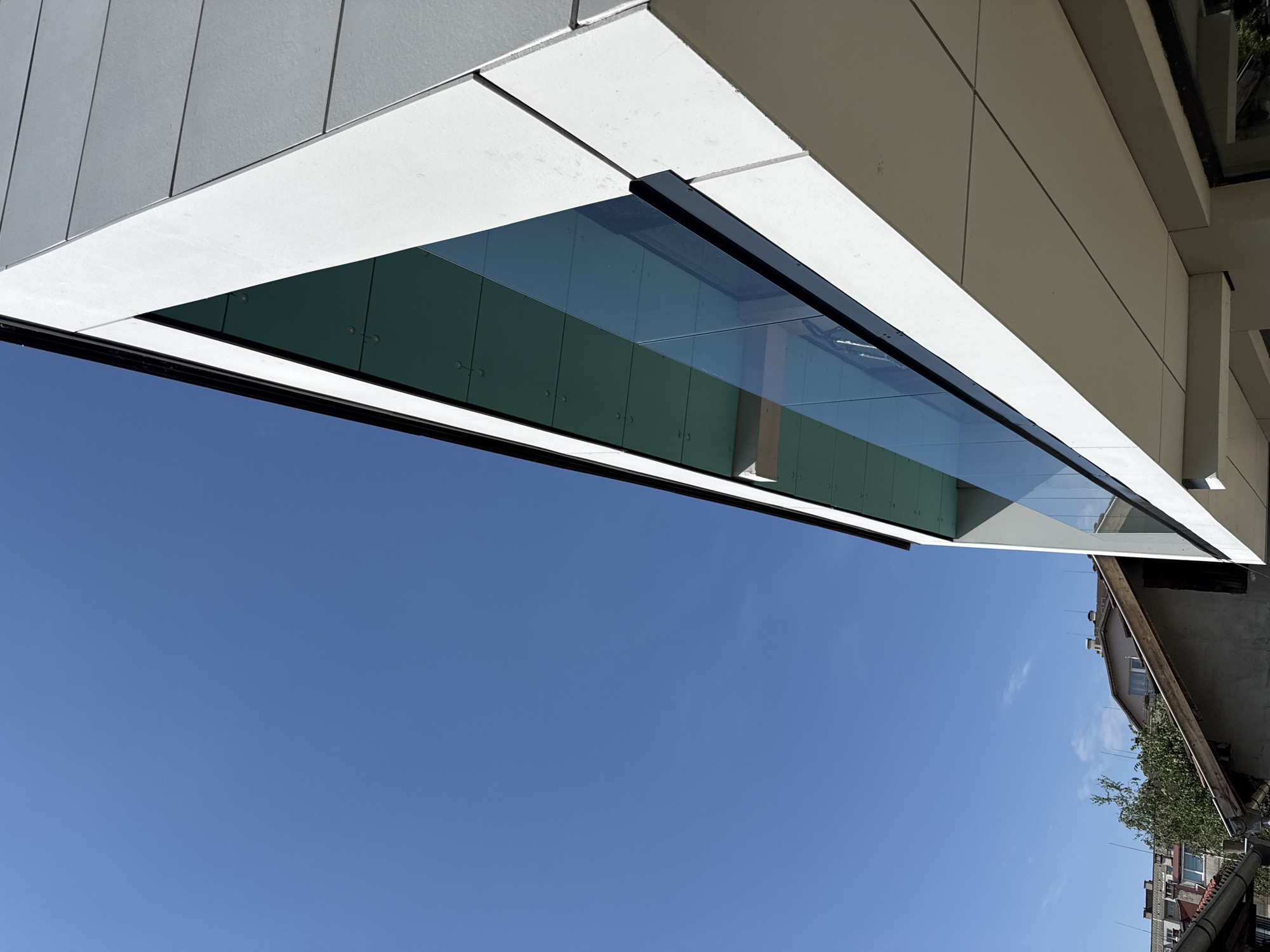 The image is a low-angle shot of a modern building with a white facade and a glass balcony, set against a clear blue sky. The building features geometric lines and appears to be constructed with large, light-colored panels. The balcony has a transparent glass railing. In the background, older buildings with tiled roofs and trees are visible, suggesting an urban environment. The image was taken in the city of Sofia, Bulgaria.