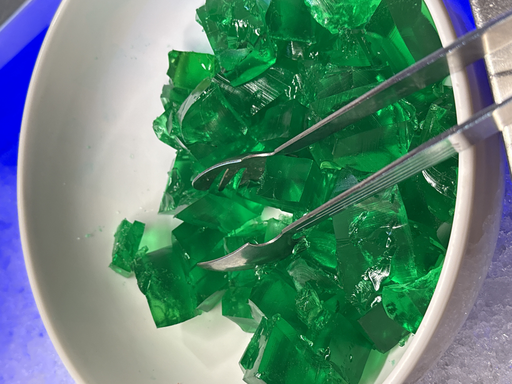 A white bowl filled with diced green gelatin is positioned on ice. Two metal serving tongs are placed in the bowl, partially submerged in the gelatin. The bowl sits in an icy display, illuminated by a blue light source from above, which also casts a glow on the top edge of the bowl. The background is blurred blue. This image was taken in Nordhausen, Germany.
