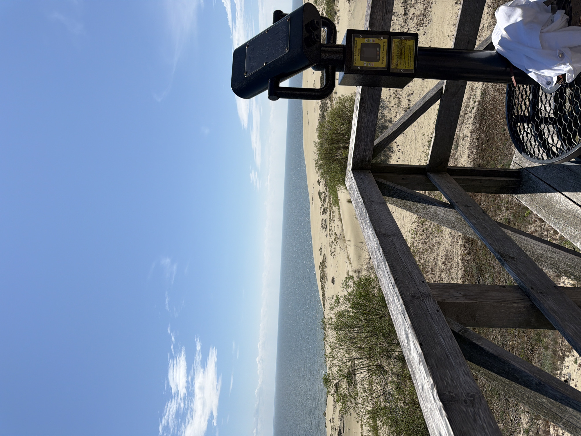 The image depicts a scenic view of the Baltic Sea and sand dunes from a wooden observation deck. In the foreground, a coin-operated viewer is mounted on a black pole, with a white shirt draped over a nearby metal grate. The observation deck is made of weathered wooden planks and railings. Beyond the deck, vast sand dunes stretch towards the horizon, dotted with sparse vegetation. The sea is calm and blue, meeting the sandy shore under a clear sky with scattered white clouds. The location is the Curonian Spit in Morskoye, Russia, known for its unique landscape of shifting sands and coastal scenery.