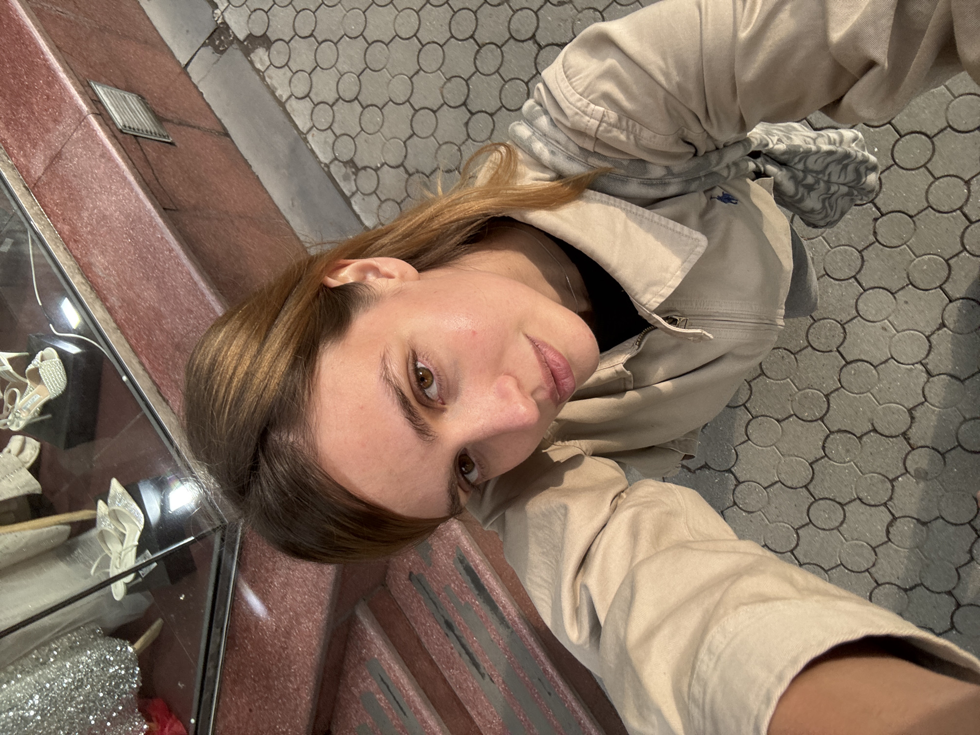 A woman is positioned above the viewer, looking down at the camera. She has brown hair, fair skin, and brown eyes. She is wearing a light tan jacket. Behind her is a sidewalk with a patterned brick design. To her left is a shop window displaying shoes. The location is Yerevan, Armenia.