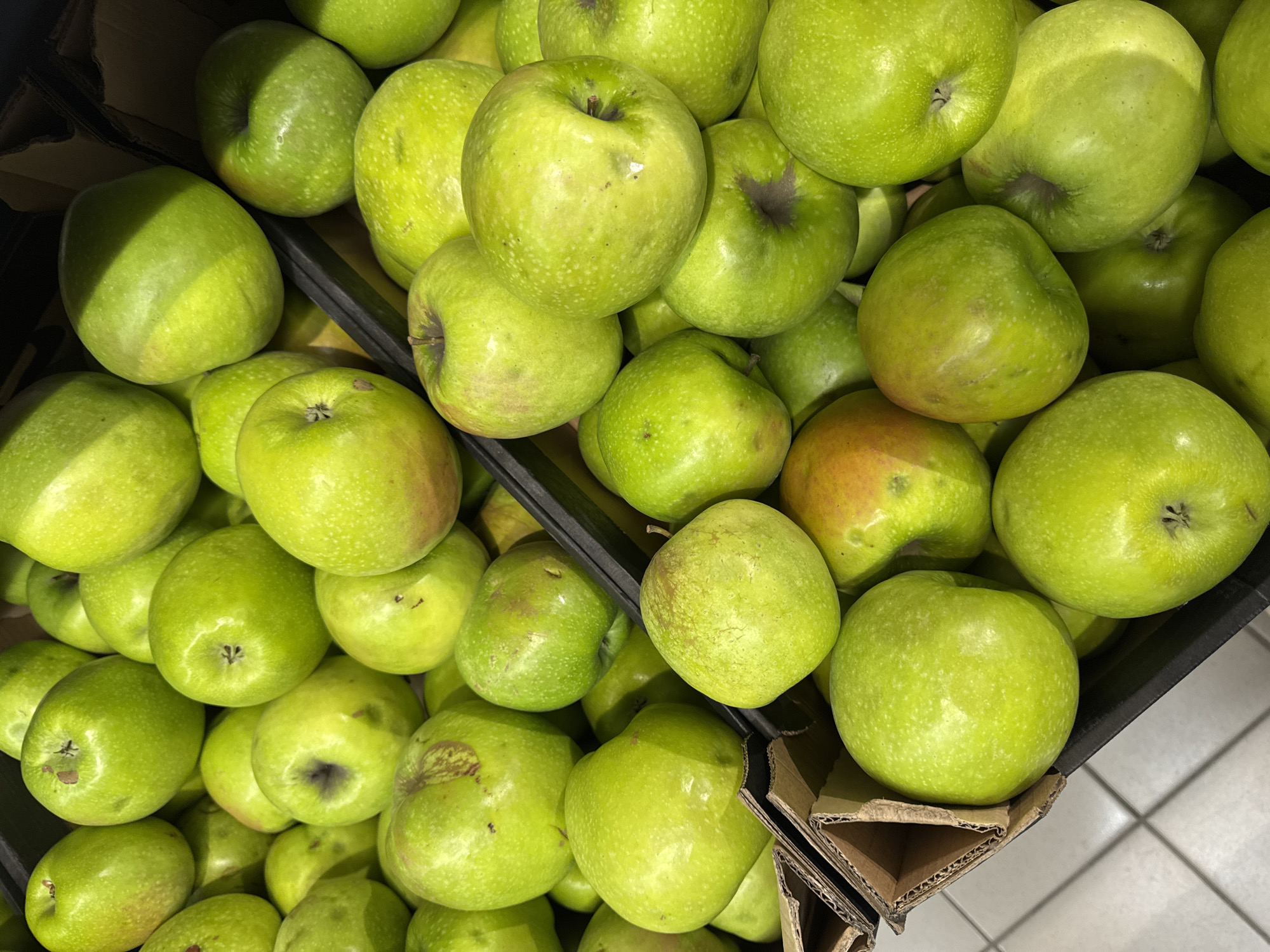 The image shows a close-up, overhead view of a display of bright green apples in a market. The apples are piled high in black plastic crates and cardboard containers. Some of the apples have small blemishes or reddish tinges. The background consists of a tiled floor and a dark, blurry area, suggesting an indoor market setting. There are no people or actions depicted in the image, only the apples themselves.