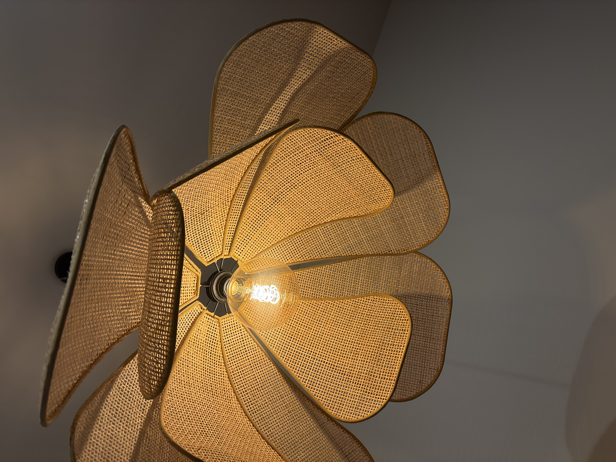 The image shows a close-up, low-angle view of a woven, flower-shaped ceiling lamp fixture with a glowing light bulb in the center. The fixture appears to be made of woven rattan or a similar material, forming overlapping petal-like structures. The light bulb is visible in the middle, emitting a warm, orange glow. The ceiling around the fixture is visible as a gradient of greys from light to dark. There is no information to indicate this image was taken in the city of Krasnoyarsk, Russia.