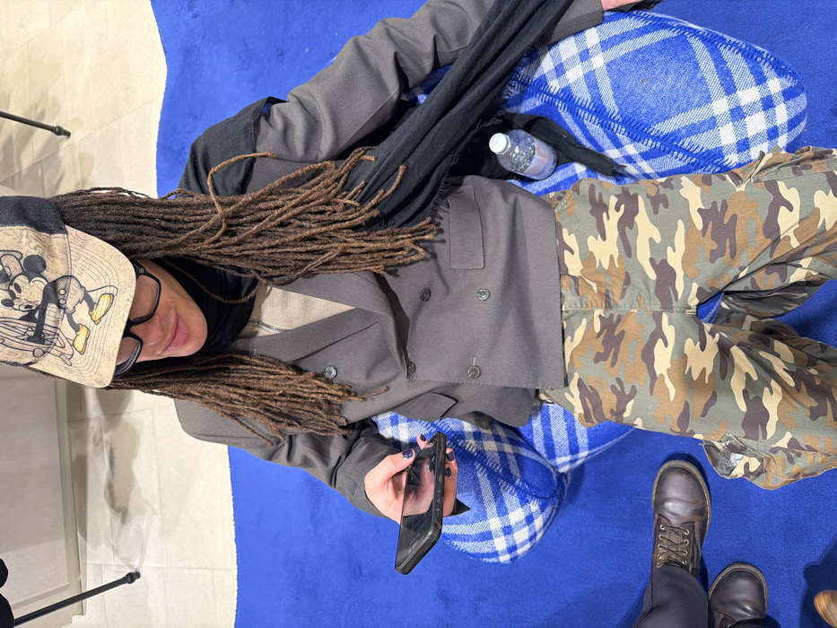 A person with dreadlocks and glasses, wearing a Mickey Mouse hat, lies on a blue surface with a blue and white checkered pillow. They are dressed in a gray blazer, a black scarf, and camouflage pants. The person holds a black smartphone. A bottle of water is nearby. Part of another person's legs and boots are visible at the edge of the frame. A microphone stand is partially visible.