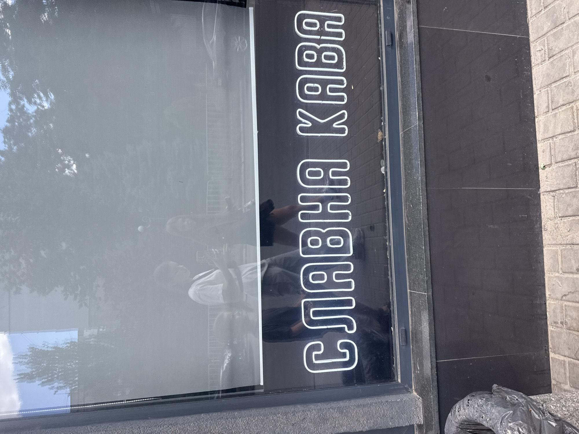 The image shows a store window in Dnipro, Ukraine, with the words "СЛАВНА КАВА" ("Slavna Kava" which translates to "Glorious Coffee") written in neon letters on the inside. The storefront appears to be at street level, with a gray tiled facade below the window and a sidewalk paved with gray bricks in the foreground. The window itself is large and reflects the exterior environment, showing blurred images of trees, buildings, and people walking by. Two individuals are visible in the reflection, one wearing a white t-shirt and the other a dark t-shirt. A black trash receptacle is situated to the left of the frame.