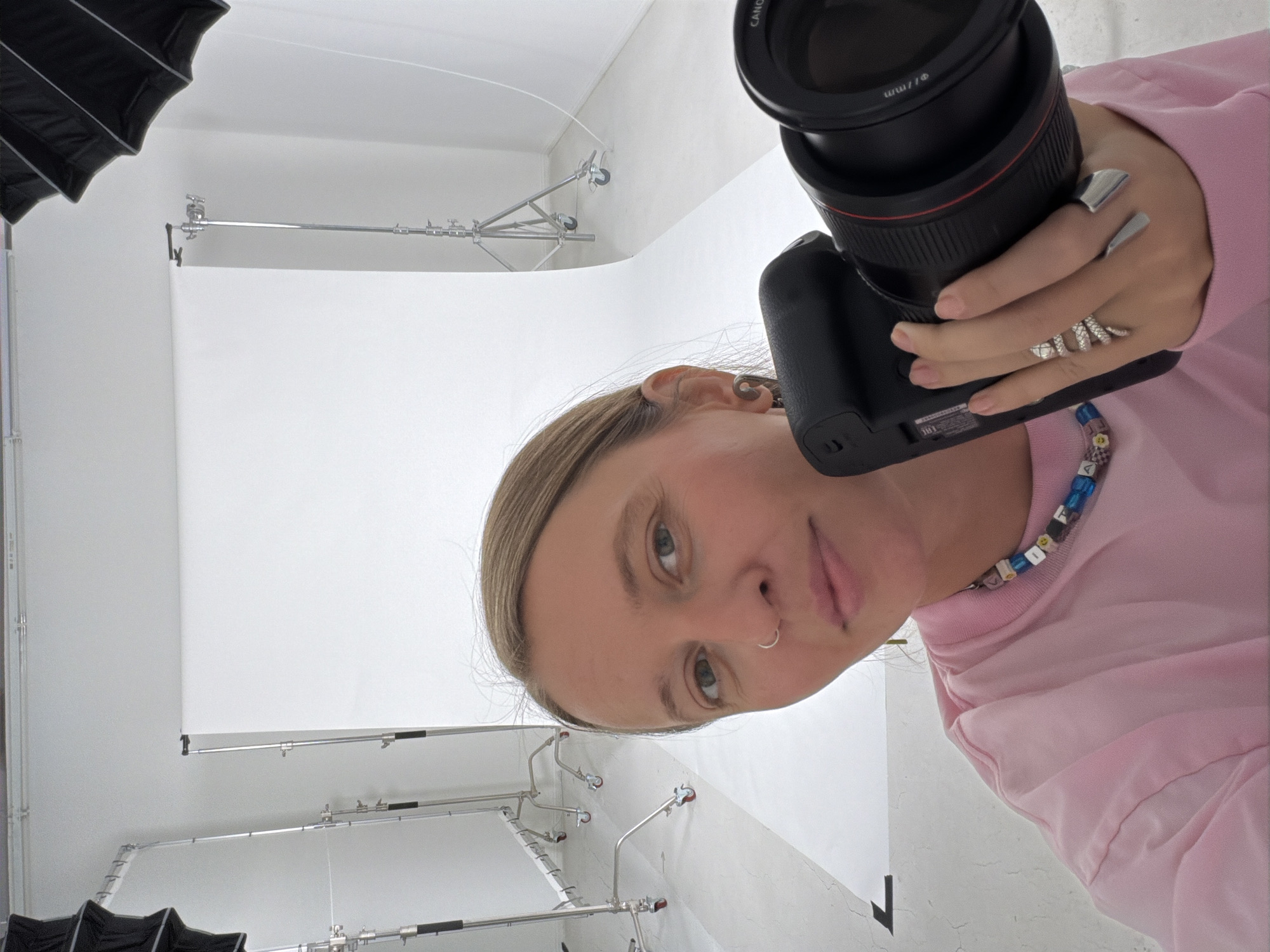 A woman is holding a camera and taking a selfie in a photography studio. The studio has a white backdrop and lighting equipment. The woman is wearing a pink shirt and a necklace with colorful beads. She is wearing several rings on her fingers. The location appears to be a photography studio in Los Angeles, United States of America.