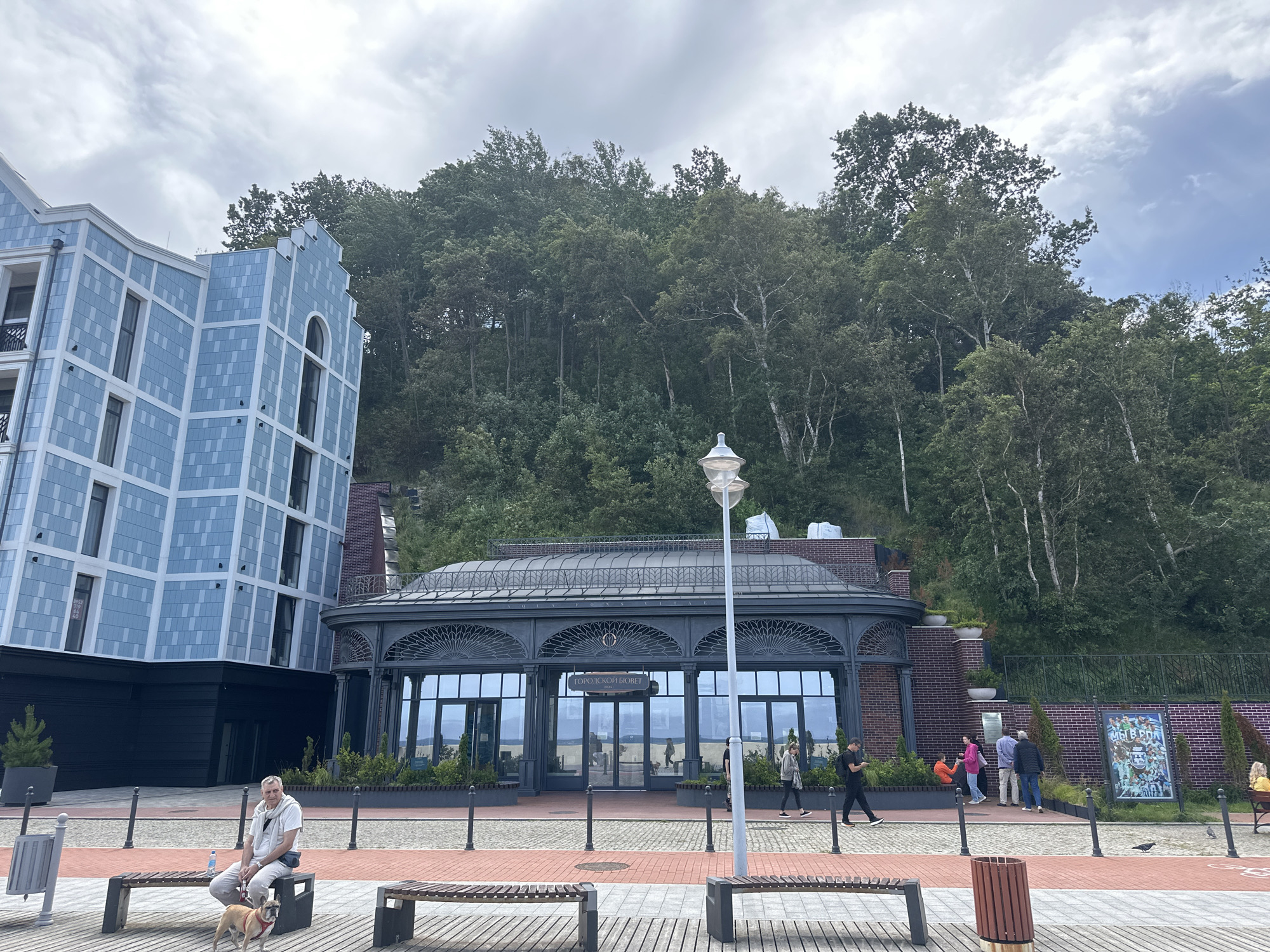 The image depicts a scene in Svetlogorsk, Russia, featuring a modern light blue apartment building with a patterned facade to the left, adjacent to a distinctive glass-enclosed pavilion with dark grey ornate metalwork. Behind these structures, a dense, lush green forest covers a steep hillside under a cloudy sky. In the foreground, a paved promenade with wooden benches runs horizontally. A man in light-colored clothing is seated on a bench on the left, with a small dog beside him. Several people are visible walking along the promenade and near the pavilion, some entering or exiting the building which has "ГОРОДСКОЙ БИОБУВЕТ" written above its entrance. Benches, trash receptacles, and lampposts are placed along the promenade, indicating a public recreational area. A colorful poster is visible on the right side of the scene.