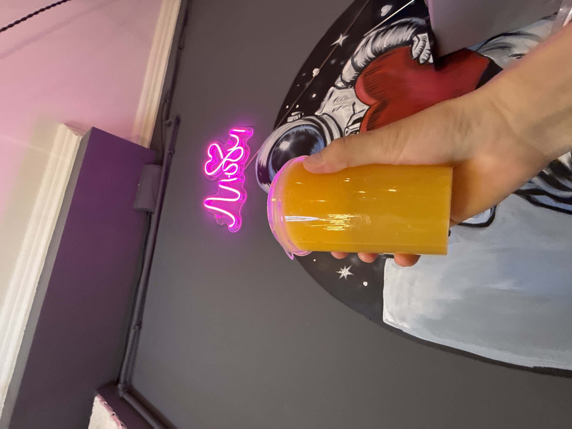 An upward angle shot shows a hand holding a clear plastic cup filled with an orange beverage. Behind the cup, a mural of an astronaut holding a large red heart is visible on a dark grey wall. Above the mural, a neon sign glows in bright pink, spelling out "LOVE." The upper portion of the image reveals a white cornice and a purple-painted ceiling. The scene appears to be indoors, possibly in a cafe or a public space, with the overall lighting characterized by the neon sign and potentially ambient indoor lighting. There is no discernible indication of the specific location in Moscow, Russia, or any specific time of day.