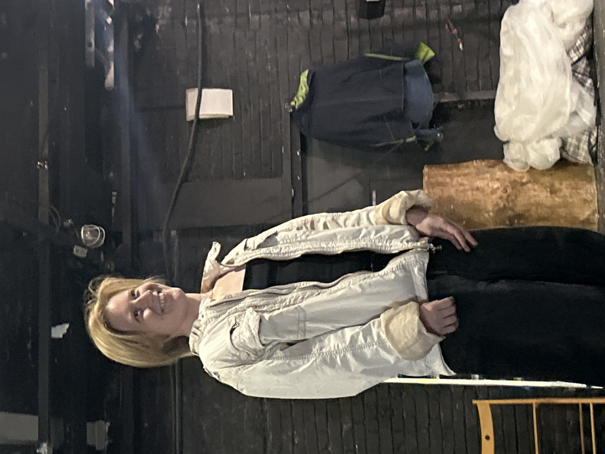 A medium shot captures a blonde woman, smiling and looking to her right, standing in an indoor, dimly lit space. She is wearing a light-colored, oversized jacket with fur-lined cuffs and a dark V-neck top underneath. Her dark pants appear to be wide-legged. To her right, a dark blue jacket with green trim hangs on a rack against a brick wall. In the foreground, a large, rough-hewn log stands upright, and to the right of that, a heap of white and plaid fabric is piled on the floor. A wooden chair is visible on the far left of the frame. The background is dark, with exposed ceiling elements and industrial lighting fixtures.