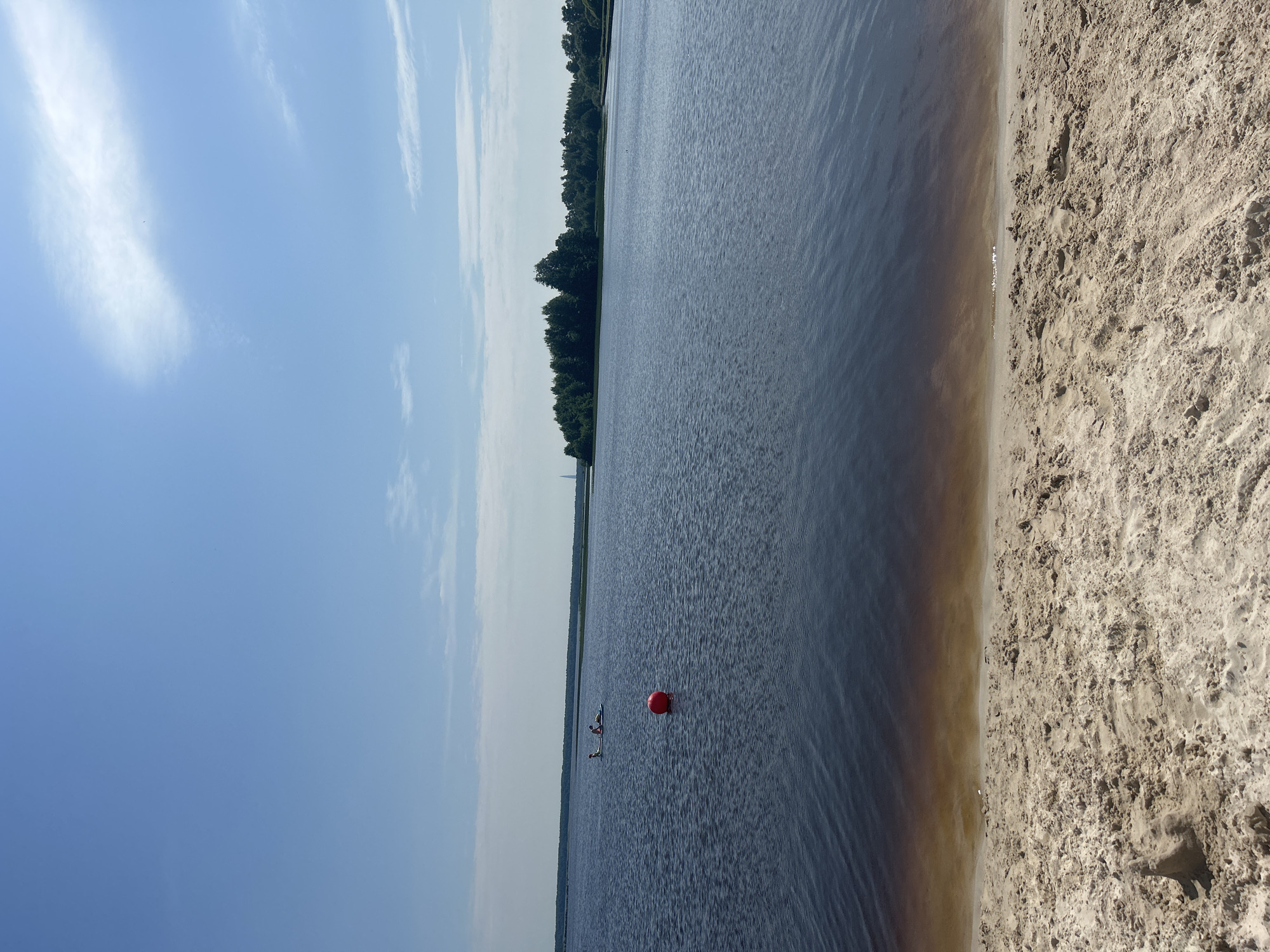 The image depicts a serene waterfront scene on a sunny day in Sestroretsk, Russia. In the foreground, a sandy beach is visible, with the gentle lapping of water at its edge. The water itself is a deep blue, with ripples reflecting the sky. Further out, a bright red buoy floats on the water, serving as a marker. In the mid-ground, two individuals are visible on a paddleboard, enjoying the water. The horizon is marked by a line of lush green trees, suggesting a natural landscape. The sky above is a clear, vibrant blue, with a few wispy white clouds scattered across it. The overall impression is one of a peaceful and picturesque summer day.