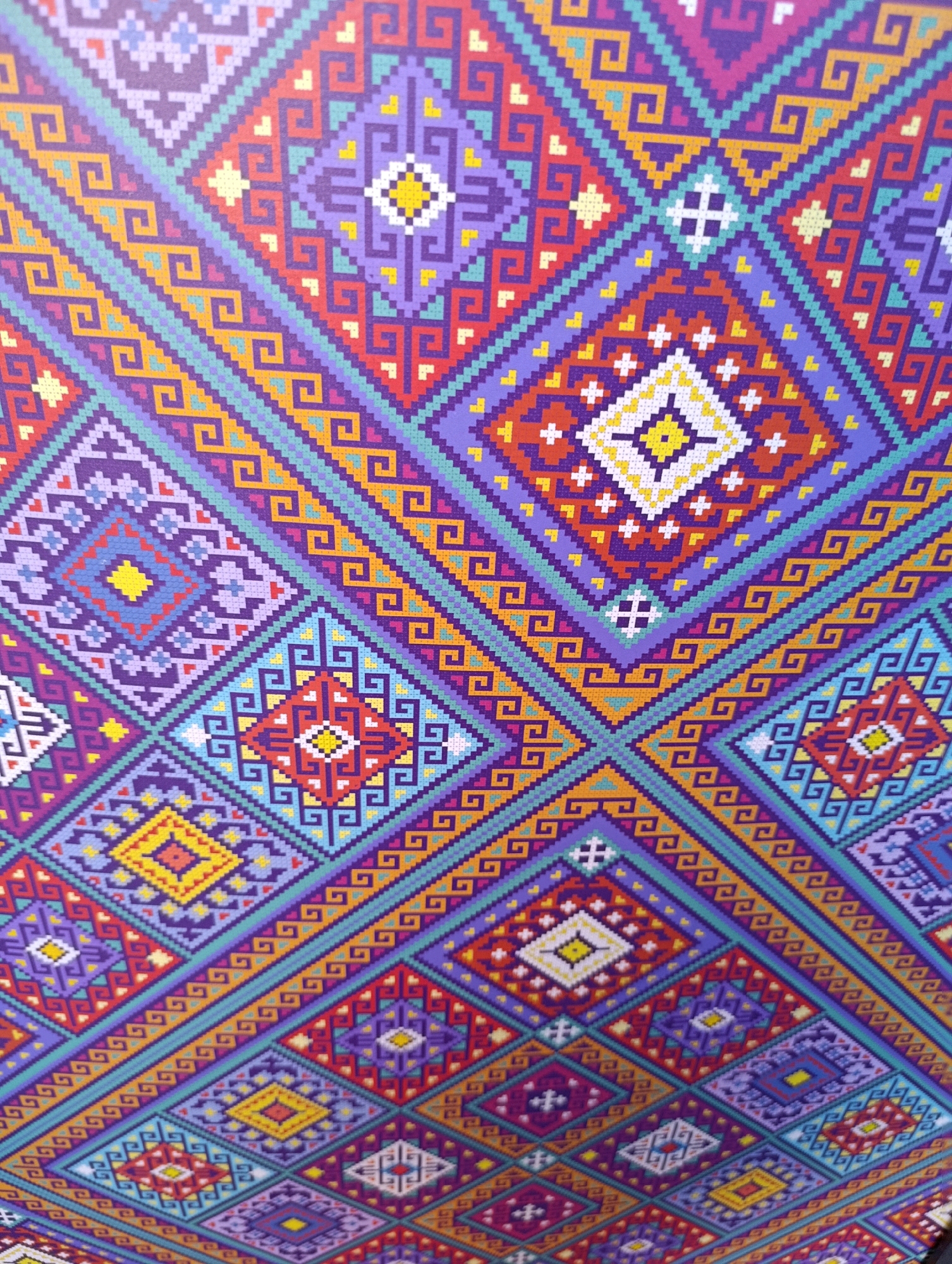 The image displays a close-up, angled view of a vibrant, geometric textile pattern. The design is intricate and appears to be woven or embroidered, featuring a repeating motif of diamonds, squares, and zig-zag lines. The color palette is rich, dominated by shades of purple, red, orange, yellow, teal, and white. The pattern is dense and highly detailed, with each element composed of small, pixel-like squares, suggesting a cross-stitch or similar technique. There are no people, identifiable landmarks, or environmental details visible that would allow for the identification of its location in Cherkasy, Ukraine, or any other specific place. The image focuses solely on the detailed visual texture and design of the fabric.