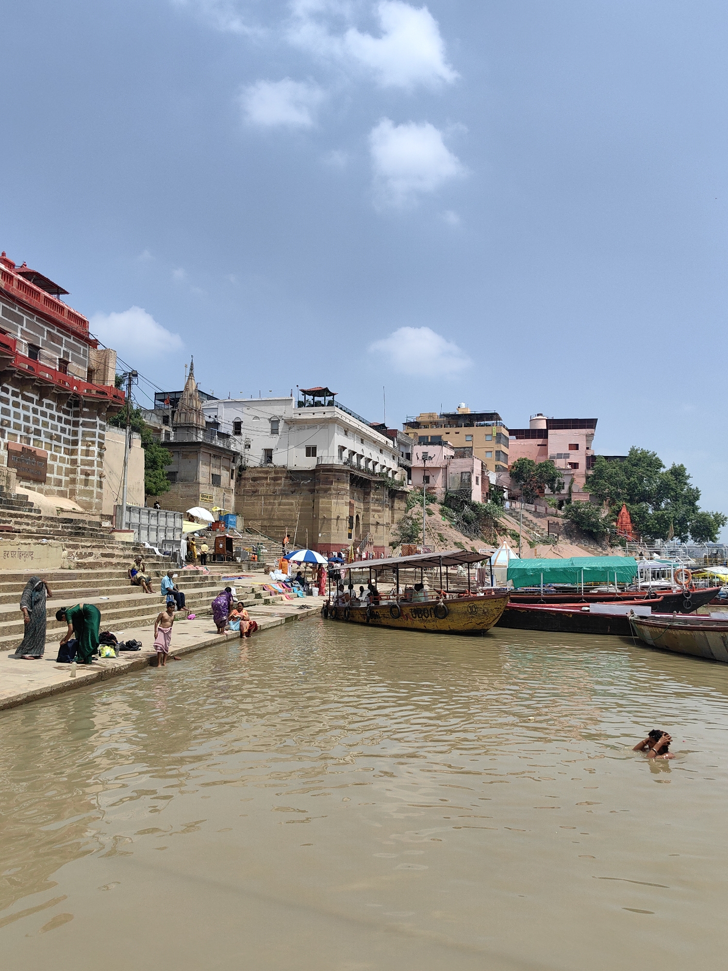 This is a photograph of the Varanasi ghats, a series of steps leading down to the Ganges River in Varanasi, India. The image depicts a bustling scene with people engaged in various activities. In the foreground, the muddy brown water of the Ganges reflects the sky. A man is swimming in the river, his head and shoulders visible above the water. On the ghats, numerous people are gathered. Some are sitting on the steps, while others are standing near the water's edge. Several women in traditional Indian attire are washing clothes and bathing. A group of people are gathered around a small stall with umbrellas.
In the background, several boats are docked along the river, including a yellow passenger boat and several other vessels. Behind the ghats, a row of multi-story buildings lines the embankment, displaying a mix of architectural styles. The buildings are painted in various colors, including pink, white, and cream. Some of these buildings appear to be temples or religious structures, with intricate carvings and spires. Green trees are visible interspersed among the buildings, adding a touch of nature to the urban landscape. The sky above is a clear blue with a few scattered white clouds. The overall atmosphere is one of daily life and religious activity taking place by the sacred river.