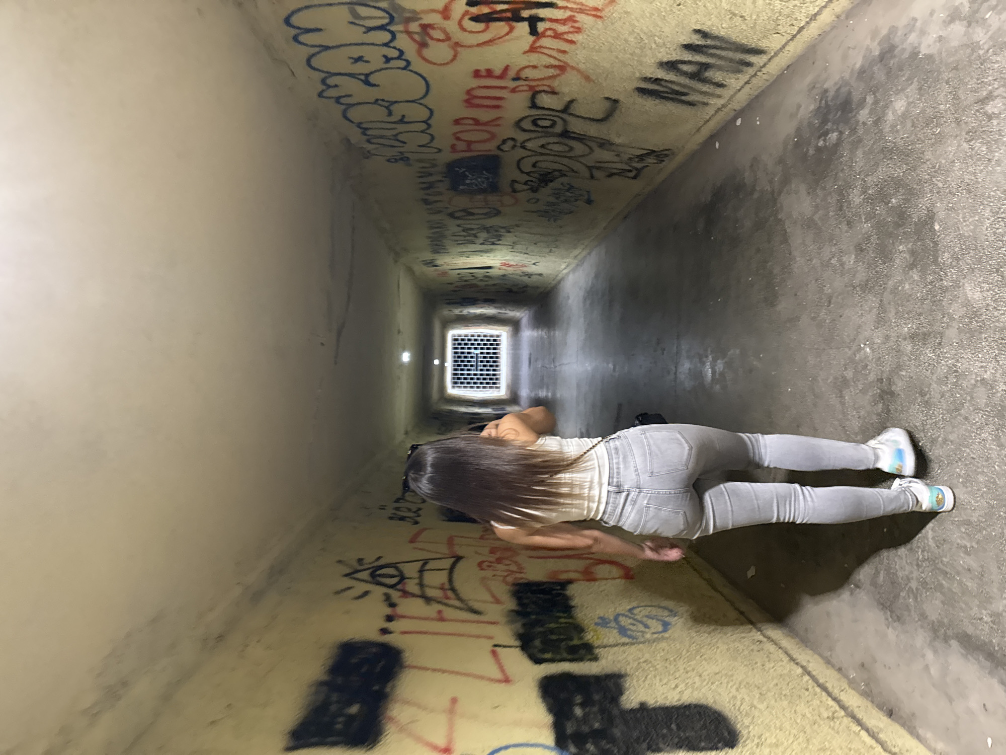 A woman with long dark hair, wearing a white top and grey jeans, is lying on her stomach in a graffiti-covered tunnel. The tunnel appears to be constructed of concrete. The graffiti is varied, including words, symbols, and drawings. The far end of the tunnel is a metal grate, illuminated with light. The specific location in Kostinbrod, Bulgaria is not evident from the image.