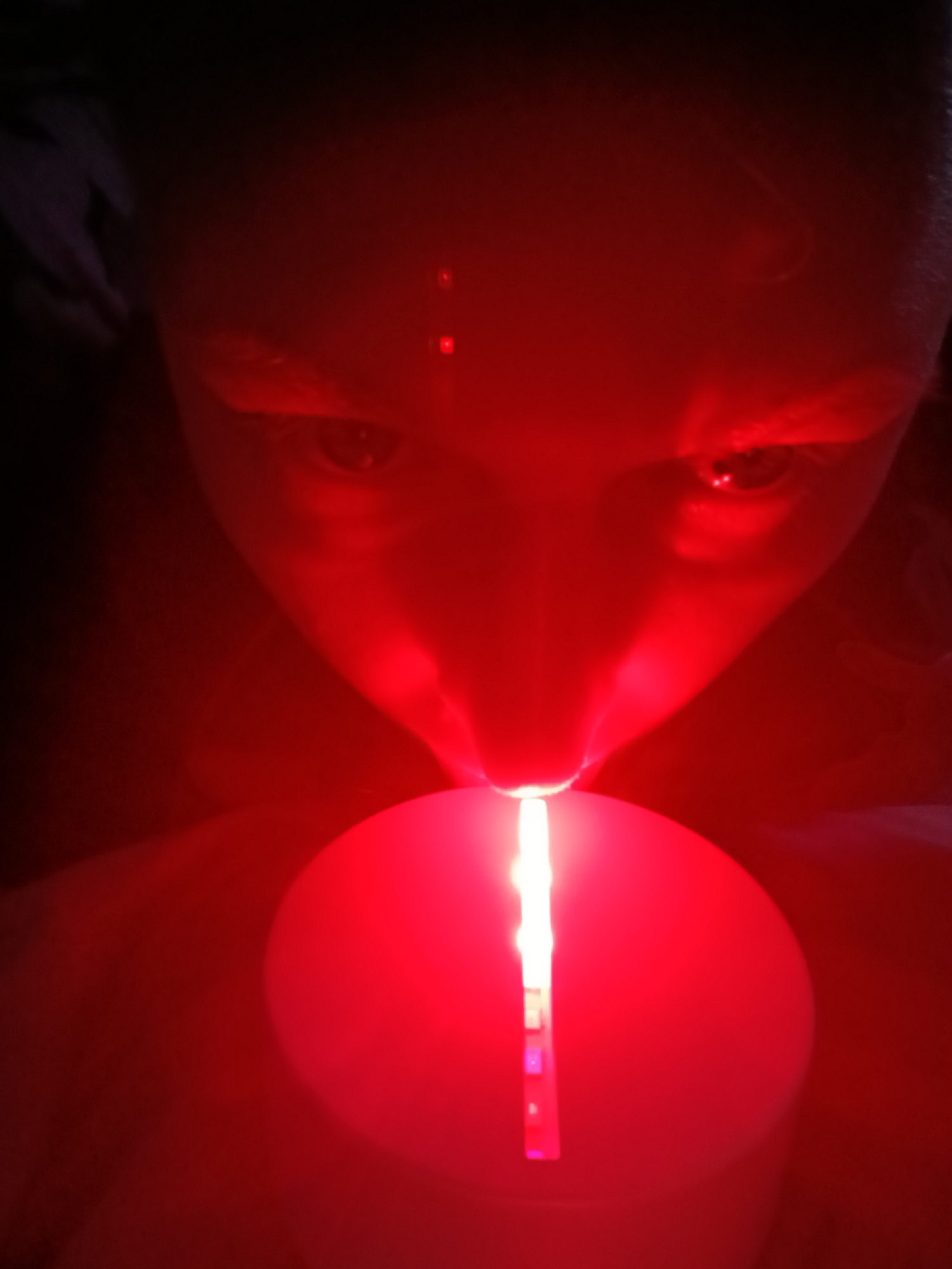 A close-up, dark image shows a person's face illuminated by a red light, likely from a lit object (possibly a candle) held below. The light shines up onto their face, creating red reflections in their eyes and on their skin. The person appears to be looking down at the light source. No specific details indicating the location in Chaikovsky, Russia, are visible.