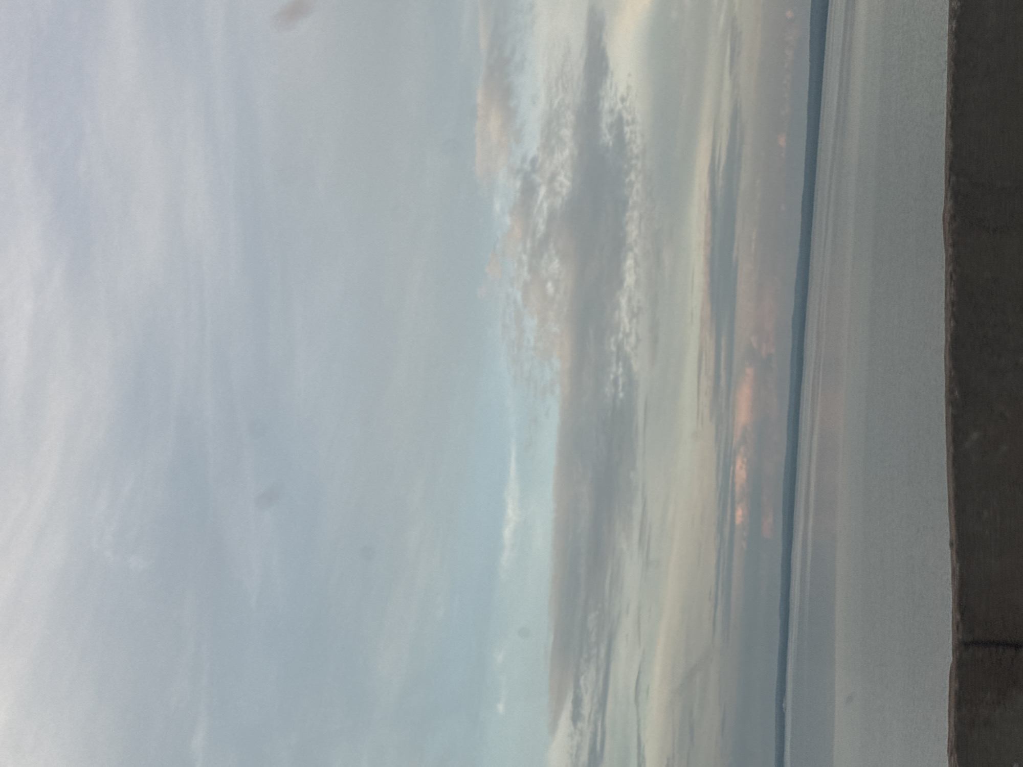 The image shows a landscape with a body of water and a cloudy sky. The water extends to the horizon, with a distant landmass visible. The sky is filled with soft, diffused clouds, with hints of pink and orange suggesting either sunrise or sunset. The overall mood is serene and tranquil. The image is oriented vertically. On the right side of the frame, a dark, textured surface, possibly a railing or a wall, is visible. There is no visible text. The time of day is likely either dawn or dusk, given the colors in the sky. The weather appears to be calm. The location context is Noviy, Russia.