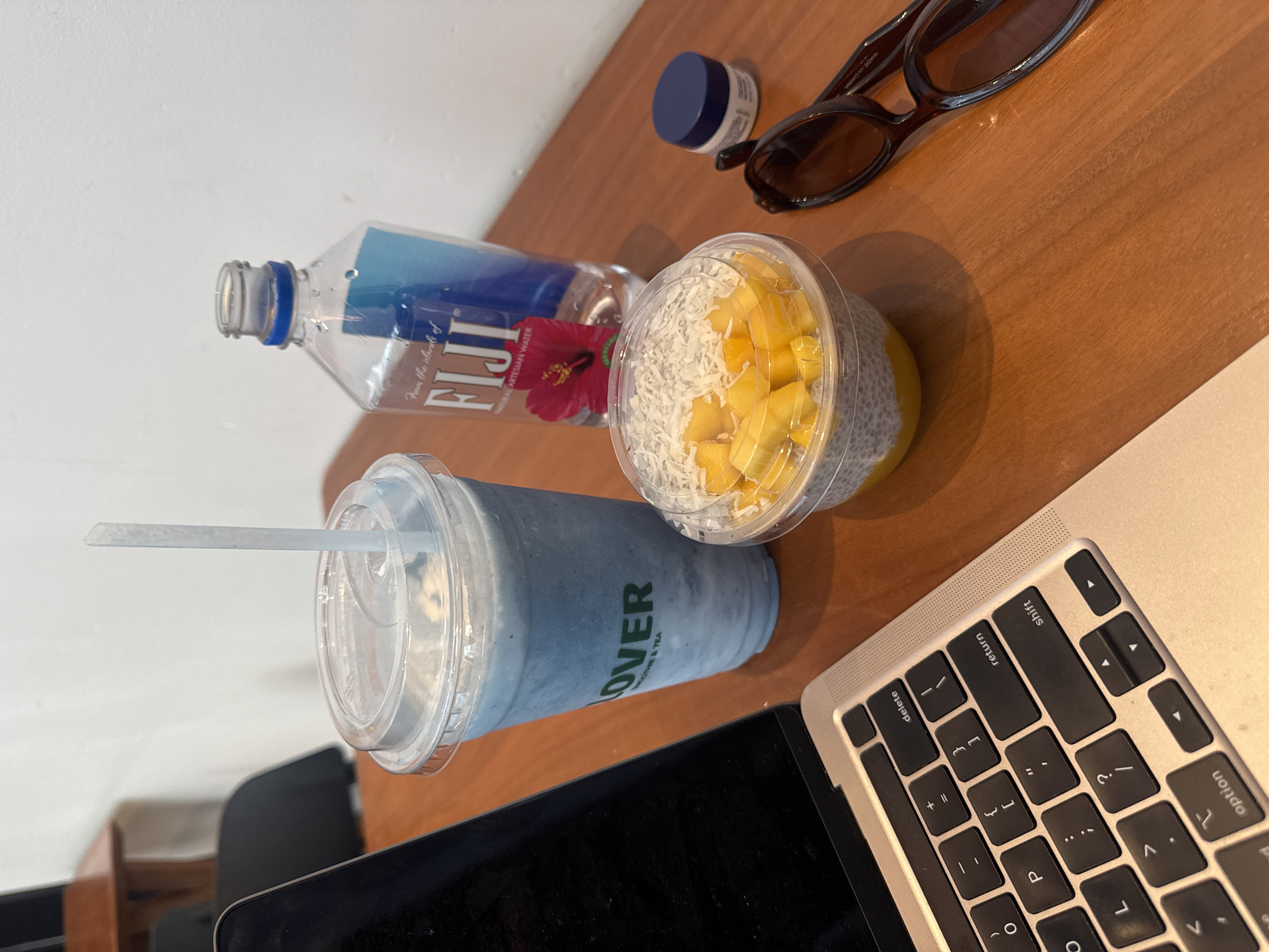 A high-angle shot captures a collection of items on a light brown wooden surface, likely a desk, with a white wall in the background, suggesting an indoor setting. The arrangement indicates a casual work or study environment, possibly in New York, United States.
Prominently featured on the left is a tall, light blue beverage cup with a clear lid and a white straw, bearing the text "OVER SMOOTHIE & TEA." To its right, a clear plastic container holds a snack resembling chia pudding, generously topped with vibrant diced mango and white shredded coconut.
Further back, a Fiji brand water bottle is visible, partially filled, with its label clearly showing "FIJI NATURAL ARTESIAN WATER" and "From the islands of," adorned with a red hibiscus flower. Adjacent to the bottle, a pair of dark sunglasses rests on the table, with "MADE IN CHINA" faintly discernible on one arm. A small, round container with a dark blue lid is also present near the sunglasses.
On the bottom right, the silver keyboard of a laptop is partially visible, displaying keys such as "delete," "return," "shift," and "option." The natural lighting casts soft shadows, indicating daylight.