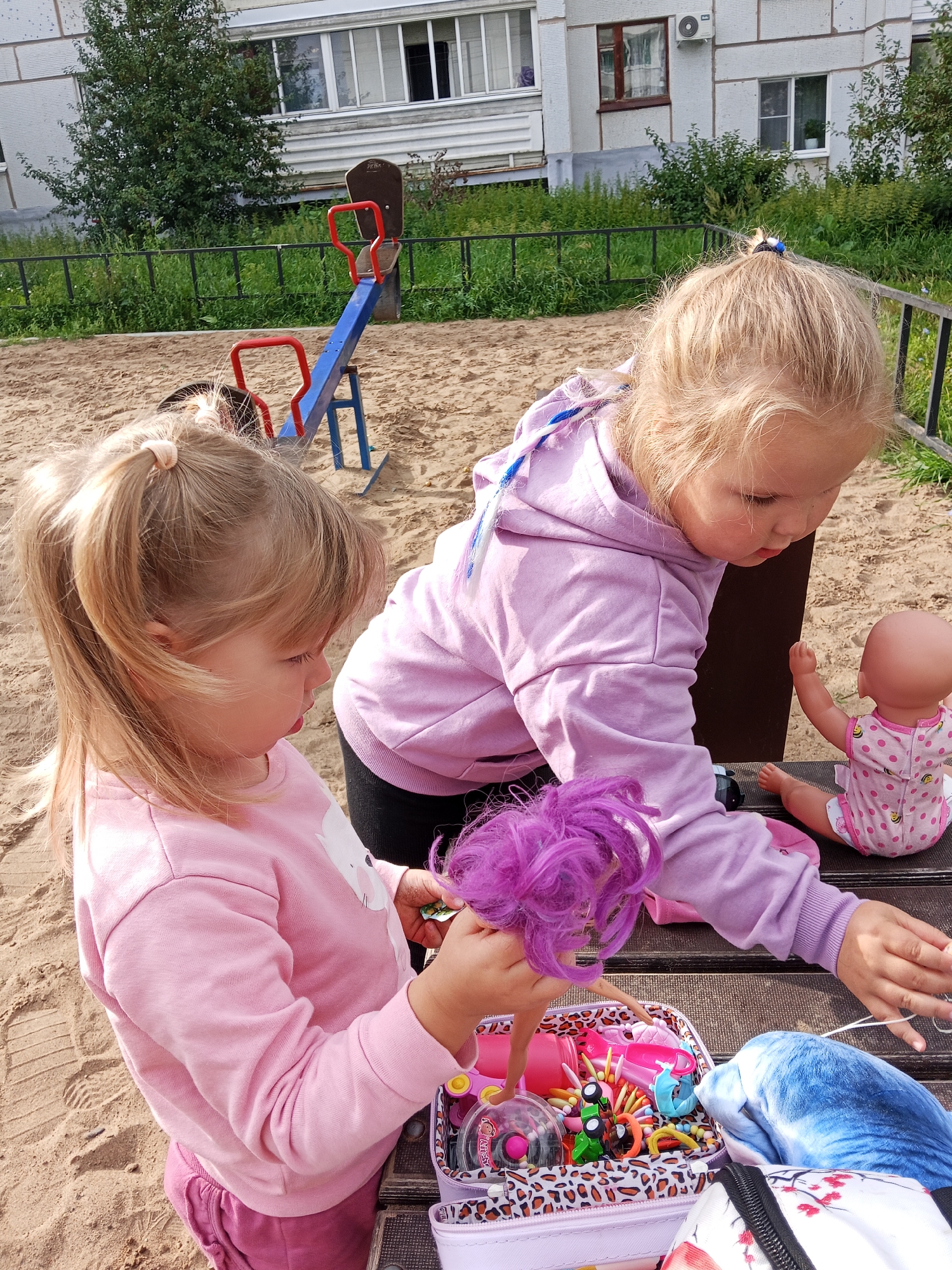 The image depicts a playground in the city of Chaikovsky, Russia, with two young girls actively engaged in play. The setting appears to be a sandbox area, with a seesaw visible in the background. The girls are dressed casually, with one wearing a pink sweatshirt and the other a purple hoodie. They are interacting with dolls and a container filled with various toys, suggesting imaginative play. The environment is outdoors, with greenery and a residential building visible behind the playground. The overall scene conveys a sense of childhood innocence and recreation in an urban setting.
