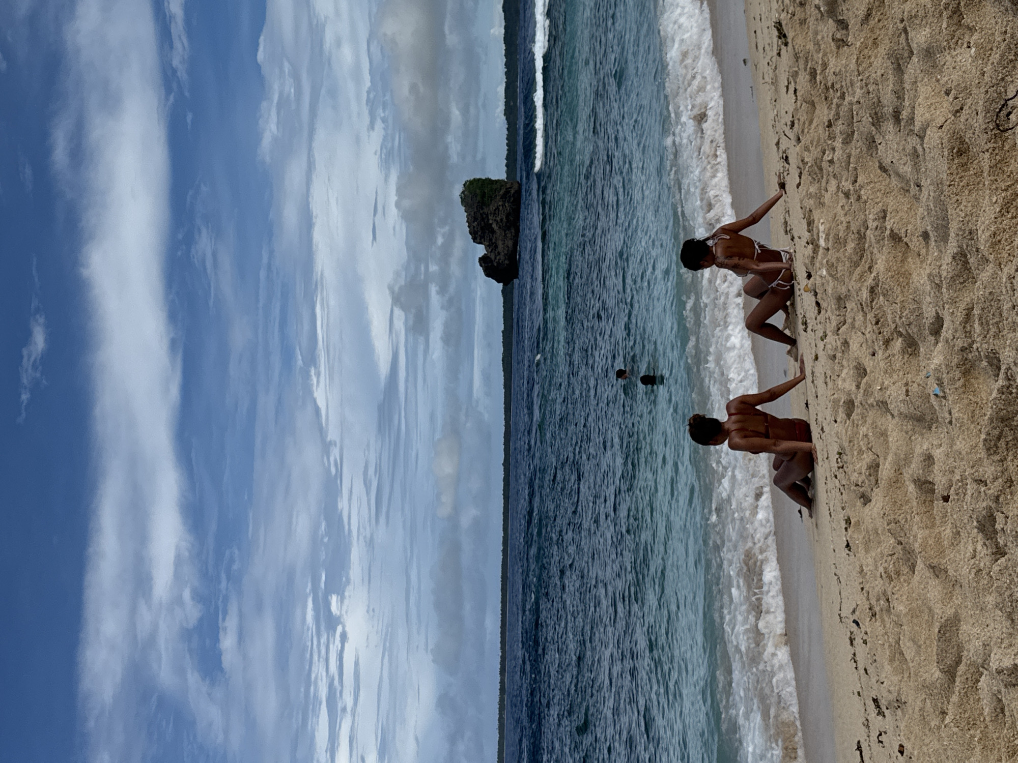 The image shows a beach scene in Pilar, Philippines. Two individuals, possibly tourists, are crouching at the edge of the shoreline. The beach is sandy with visible wave action. In the background, a rock formation is present in the water. Further out, other people can be seen in the water. The sky is blue with varied cloud formations.