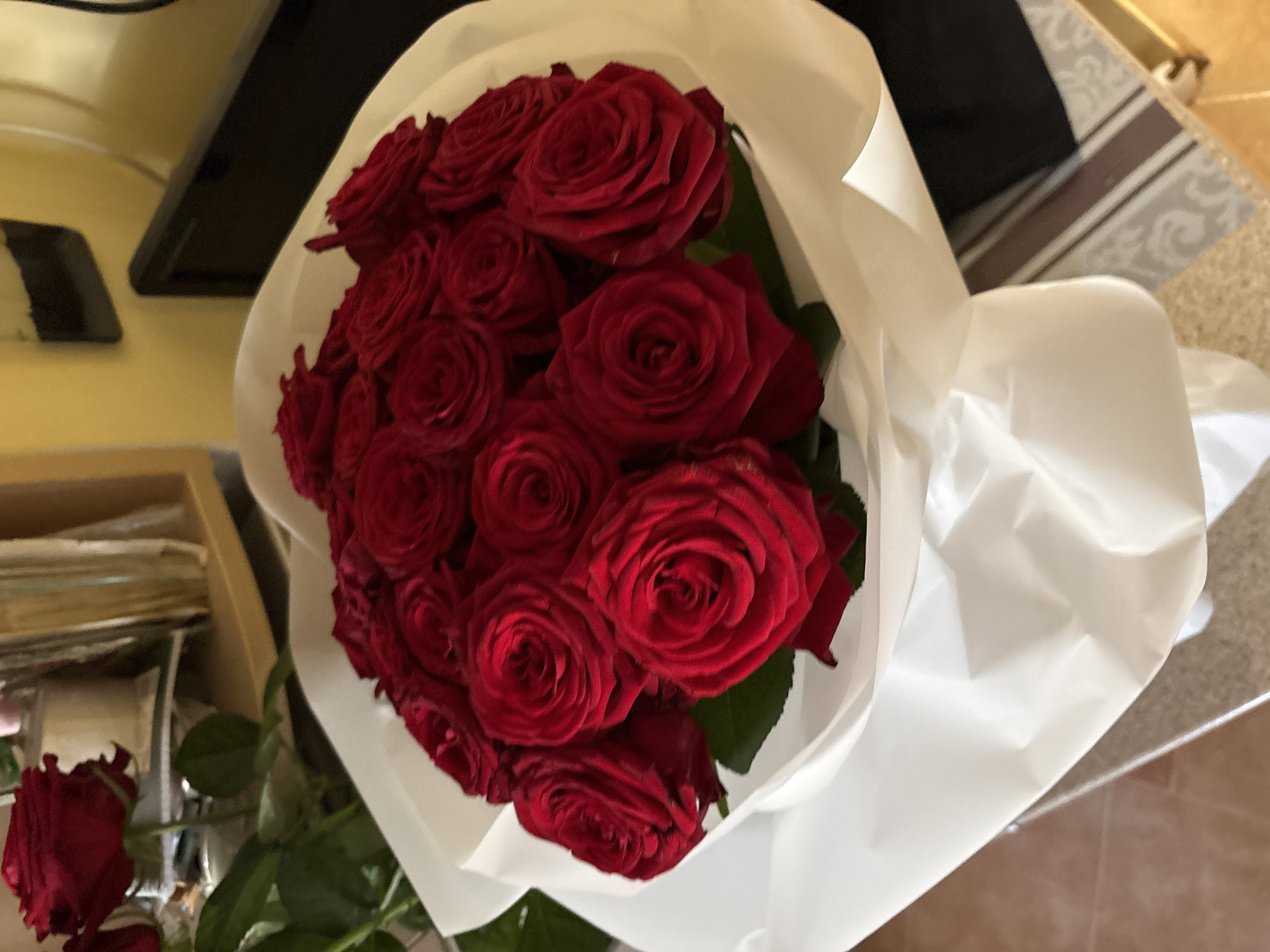 A bouquet of deep red roses is presented in a white paper wrap. The roses are tightly packed and their velvety petals are visible. Some green leaves are interspersed amongst the blooms. The bouquet is resting on a surface, possibly a table, with a tiled floor visible in the lower left corner. In the background, a yellow wall and a dark object, possibly a monitor, can be seen. The image is taken from a slightly elevated angle, focusing on the roses. There is no indication of the city of Minsk, Belarus within the image.
