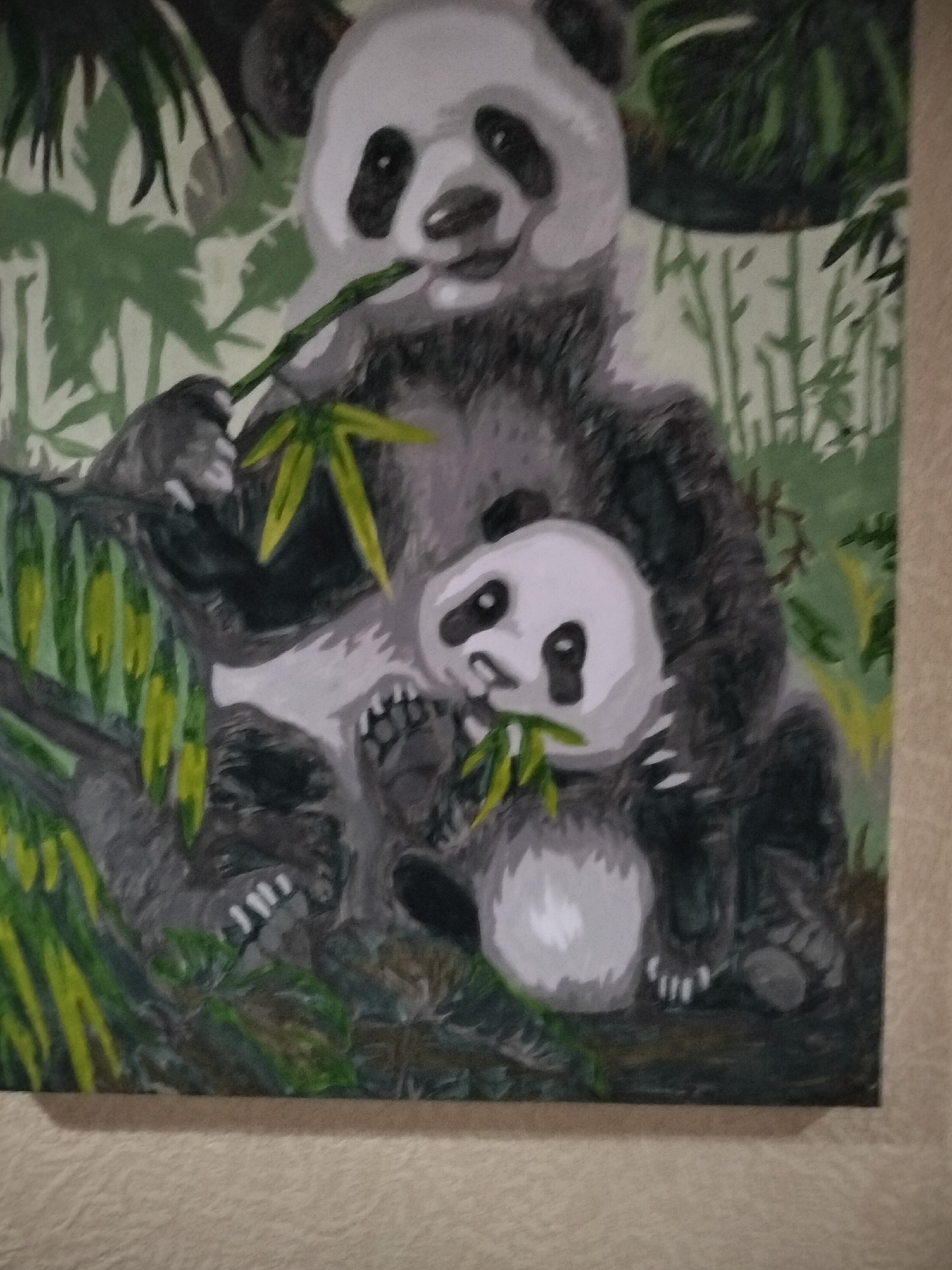 This is a painting of two pandas, one adult and one cub, nestled amongst bamboo. The adult panda is on the left, holding a stalk of bamboo to its mouth. The cub is nestled in the adult's lap, also eating bamboo. The background is a muted green, with hints of darker greens representing more bamboo. The painting is framed by a light-colored textured surface that appears to be a wall. The painting is not located in the city of Nizhnedonskoy, Russia; there is no information in the image to suggest a specific location.
