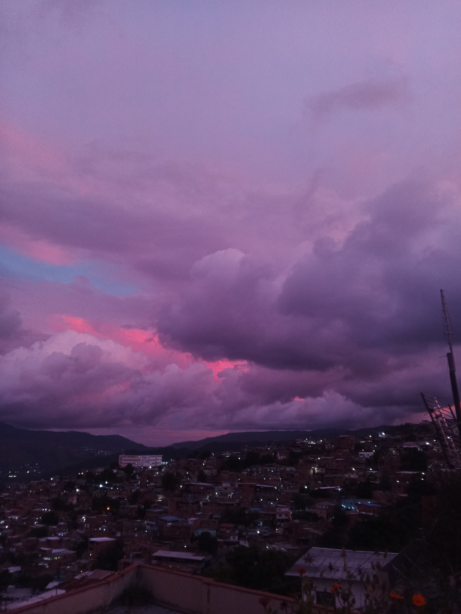 The image depicts a twilight scene over a densely populated urban area, likely a hillside barrio within Caracas, Venezuela. The sky is dominated by dramatic, voluminous clouds, tinged with vibrant shades of pink and purple, indicative of a sunset or sunrise. Patches of lighter blue are visible through the clouds, suggesting a break in the atmospheric conditions.
Below the sky, a sprawling cityscape unfolds, characterized by numerous tightly packed buildings with red-tiled roofs and brick walls, characteristic of informal settlements. Small points of light, representing illuminated windows and streetlights, dot the landscape, indicating that the city is coming alive as darkness falls. The buildings are built on uneven terrain, with many structures cascading down the hillside.
In the mid-ground, a larger, more modern building with multiple stories and windows is visible, standing out from the surrounding residential structures. The foreground is occupied by the edge of a rooftop, with a concrete structure and some vegetation. A metallic pole with a grid-like structure, possibly a communications antenna or part of a fence, is positioned on the right side of the image, partially obscuring the view of the city. The overall impression is one of a vibrant, densely populated community nestled against a backdrop of dramatic natural beauty.