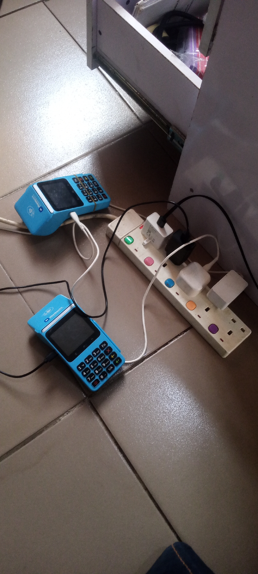 The image shows a close-up view of two blue Point of Sale (POS) terminals plugged into a power strip. The POS terminals are on a tiled floor. One POS terminal is closer to the foreground, and another is slightly further back and to the left. Both terminals are blue with black screens and keypads. White and black charging cables are connected to the POS terminals and plugged into the power strip, which is white and has multiple sockets. Some of the sockets have individual on/off buttons that are illuminated with different colors, including green, pink, orange, and purple. Multiple white and black power adapters are plugged into the power strip. In the background, a white drawer or cabinet is partially visible, with some items inside, including what appears to be plastic-wrapped items and some books. The floor is tiled with light brown square tiles. The lighting suggests an indoor environment, possibly a home or office. The location is identified as Jalingo, Nigeria.