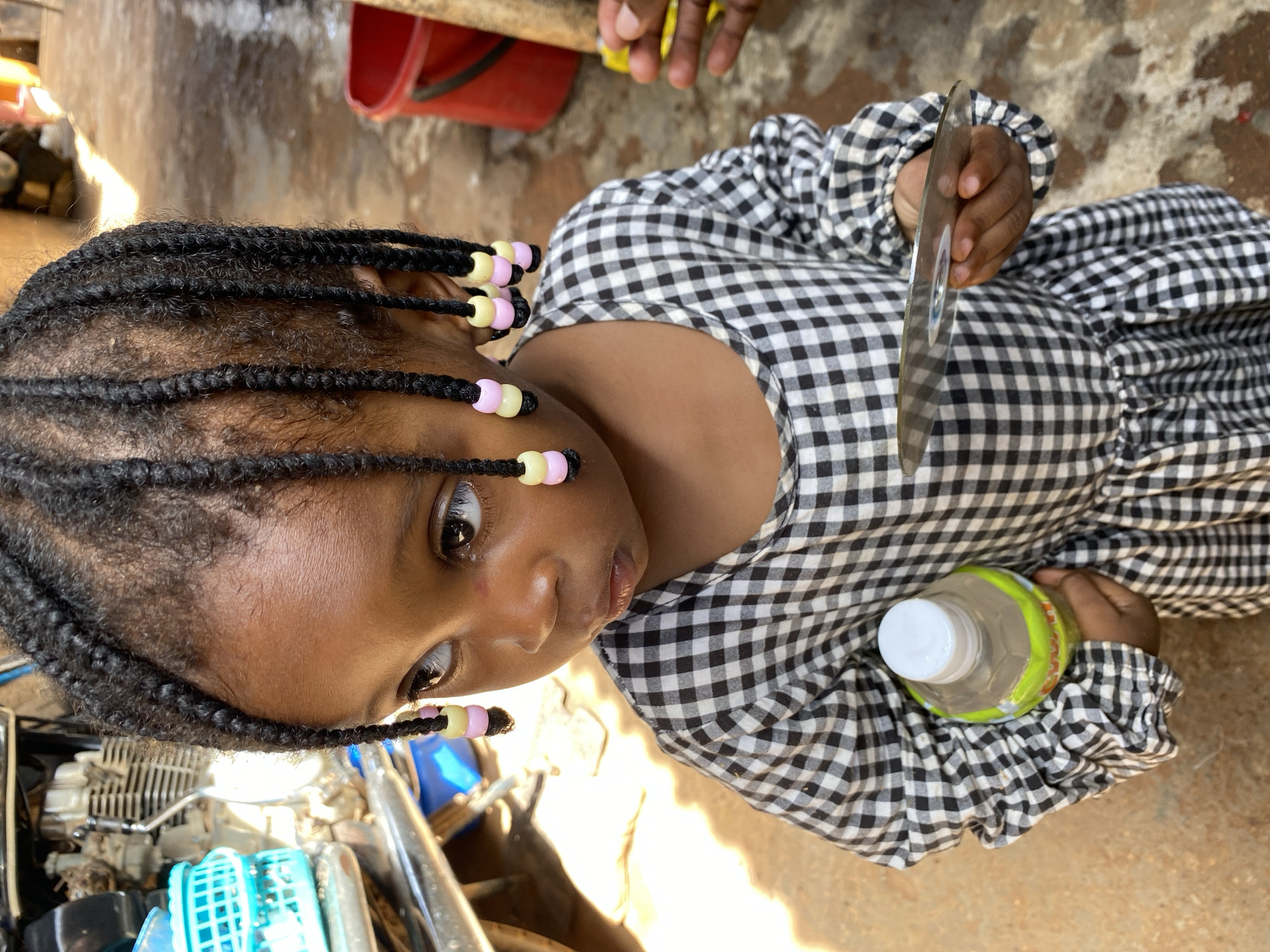 A young child with dark skin, wearing a black and white checkered dress, is the central subject of the image. The child has braided hair decorated with colorful beads in shades of pink and yellow. Their face is turned towards the viewer, with large, expressive eyes. In their right hand, the child holds a clear, flat object, possibly a CD or a piece of plastic, reflecting light. Their left arm is resting on their lap, near a clear plastic bottle with a white cap and a yellowish-green label.
The child appears to be sitting or lying down, with their body angled relative to the camera. The background is out of focus but shows a textured, earthy ground. In the upper left portion of the image, a red bucket and some metal objects are visible, suggesting an outdoor or perhaps a workshop setting. The lighting suggests it could be daytime. The overall mood seems calm, with the child exhibiting a curious or thoughtful expression. There is no visible text in the image.
