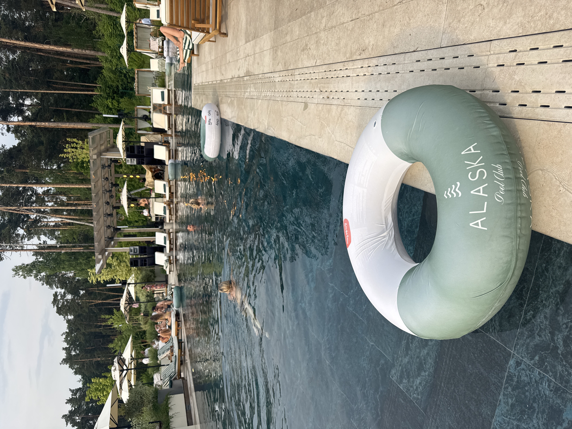The image depicts an outdoor swimming pool area, likely a resort or a club, situated in Razdory, Russia. In the foreground, a large, partially submerged inflatable ring with white and green sections rests on the pool edge. The green section bears the white lettering "ALASKA Pool Club" and a wave-like logo. The pool itself is dark blue with visible ripples.
In the background, several people are visible. Some are lounging on chairs by the poolside, while others are in the water. A woman with blonde hair is swimming in the pool. Beyond the pool, a stage with audio equipment is set up, suggesting an event or entertainment is taking place. Numerous white umbrellas are dotted around the area, providing shade. The backdrop consists of a dense forest of tall pine trees under an overcast sky. The poolside is paved with light-colored tiles and features a drainage system. Several wooden lounge chairs are arranged on the tiled area.