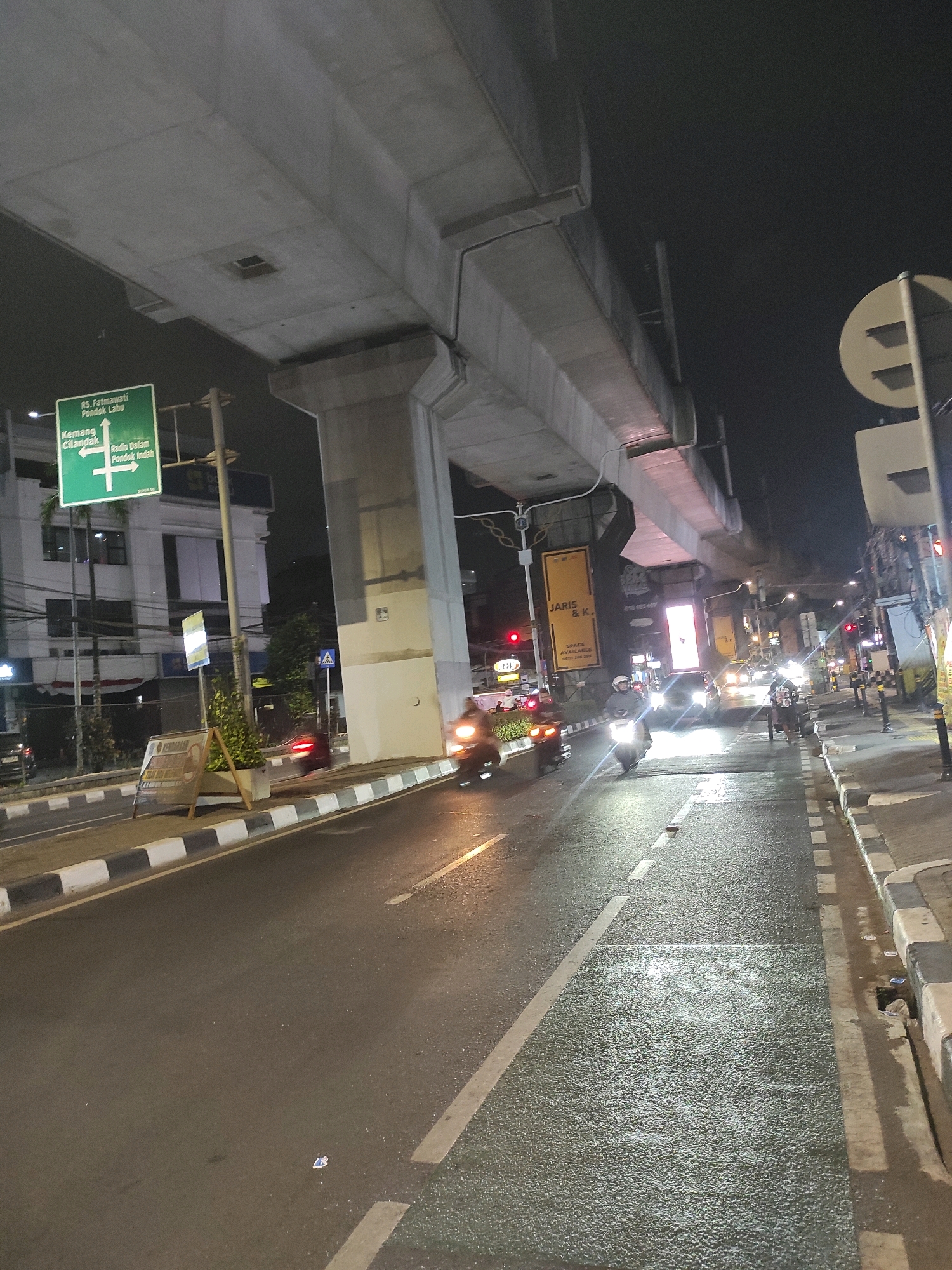 The image depicts a street scene at night in South Jakarta, Indonesia, beneath an elevated infrastructure, likely a highway or transit line. The road is wet, reflecting the streetlights and headlights of vehicles. Several motorcycles and a few cars are visible on the road, with some individuals riding the motorcycles. A traffic sign indicating directions to "RS. Fatmawati Pondok Labu," "Kemang Cilandak," and "Radio Dalam Pondok Indah" is prominently displayed on the left side of the road. Buildings and commercial signs line the street. The elevated structure casts shadows, adding to the nighttime ambiance. The overall impression is that of an urban environment with active traffic and ongoing development.