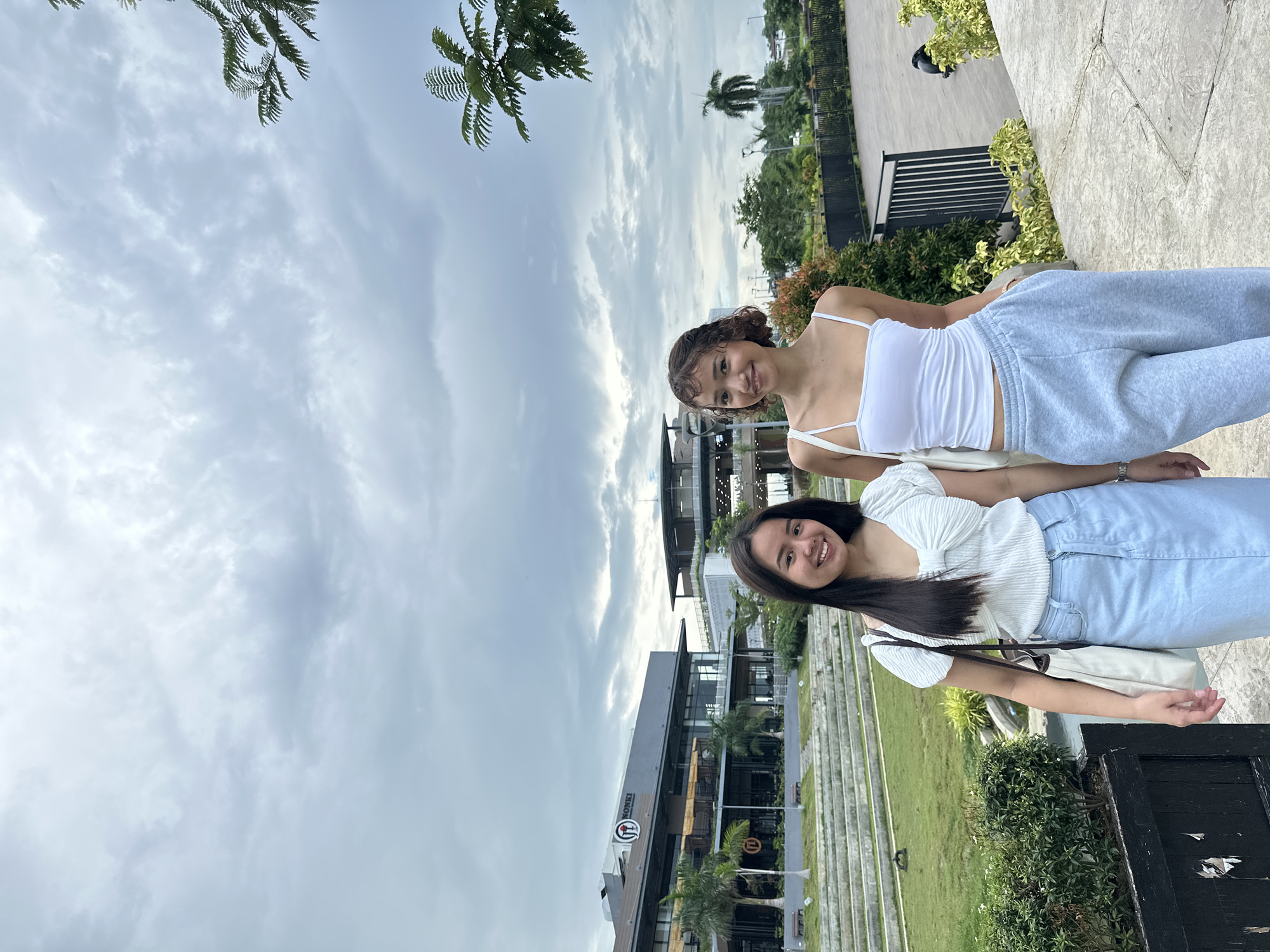 The image shows two young women standing in front of a modern building complex in Davao City, Philippines, with a cloudy sky above. The woman on the left has long, dark hair and is wearing a white textured top with a bow detail and a light blue denim skirt. She is carrying a white tote bag. The woman on the right has short, curly brown hair and is wearing a white tank top and light gray sweatpants. They are both smiling at the camera. Behind them is a tiered grassy area with concrete steps, leading up to a multi-story building with a sign that reads "NONKI." Palm trees and other greenery are visible throughout the scene. A black metal fence runs along the right side of the frame, and a black bollard is visible in the lower left corner. The overall impression is that of a casual outdoor setting in a commercial or recreational area.