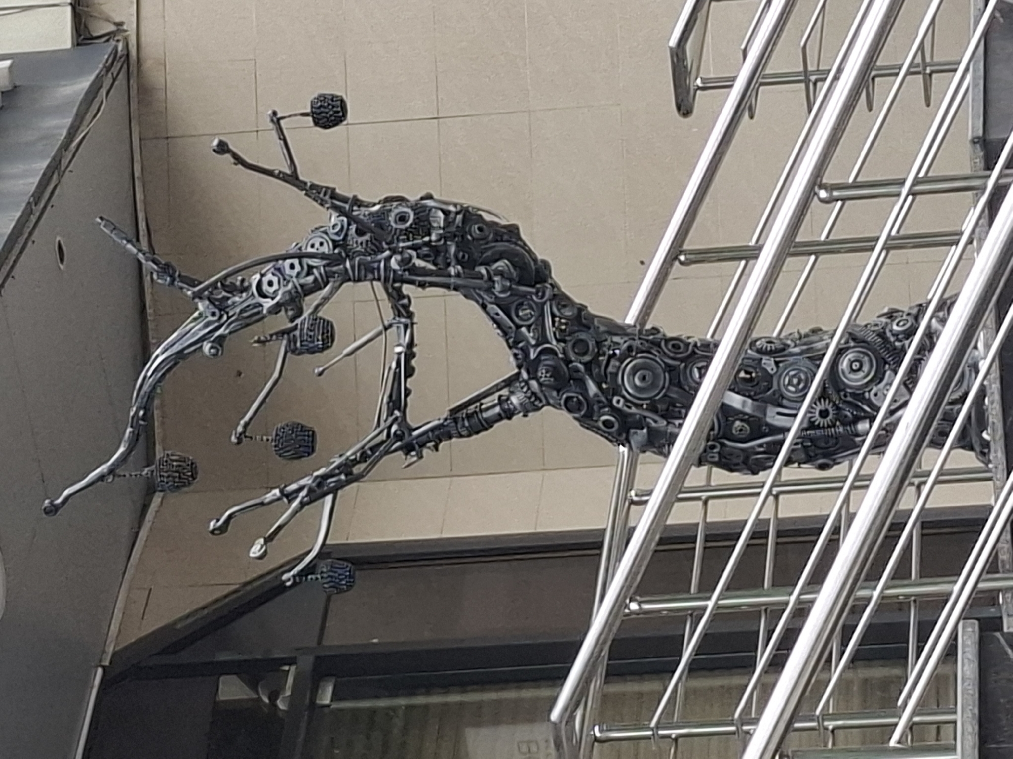 A sculpture of a tree made from metal mechanical parts, resembling gears and other industrial components, is located on an outdoor staircase. The tree has several branches, from which hang decorative elements resembling baskets or fruit, also made of metal. The staircase is lined with polished metal railings. In the background, a building with a beige tiled facade is visible, with a dark-colored entrance to the left of the sculpture. The overall scene is set in an urban environment. The sculpture is a prominent feature of the outdoor area.