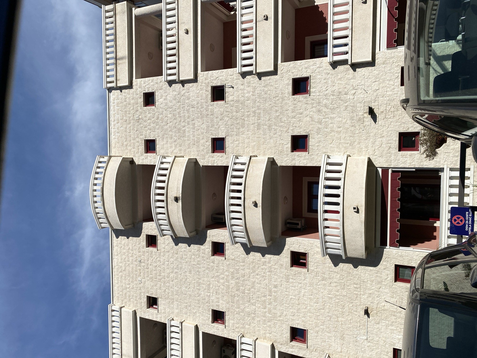 The image shows a building facade in Budva, Montenegro. The structure is multi-story, constructed with light-colored stone or concrete. It features a symmetrical arrangement of square windows with red frames. Some windows are adorned with balconies, some rectangular and some rounded. There are visible air conditioning units on some of the balconies. The sky is blue with scattered clouds. Parts of two vehicles are visible on the right side of the building. A parking sign is partially visible, attached to the building near the vehicles. The building is "Hotel Dimic Ellitt".